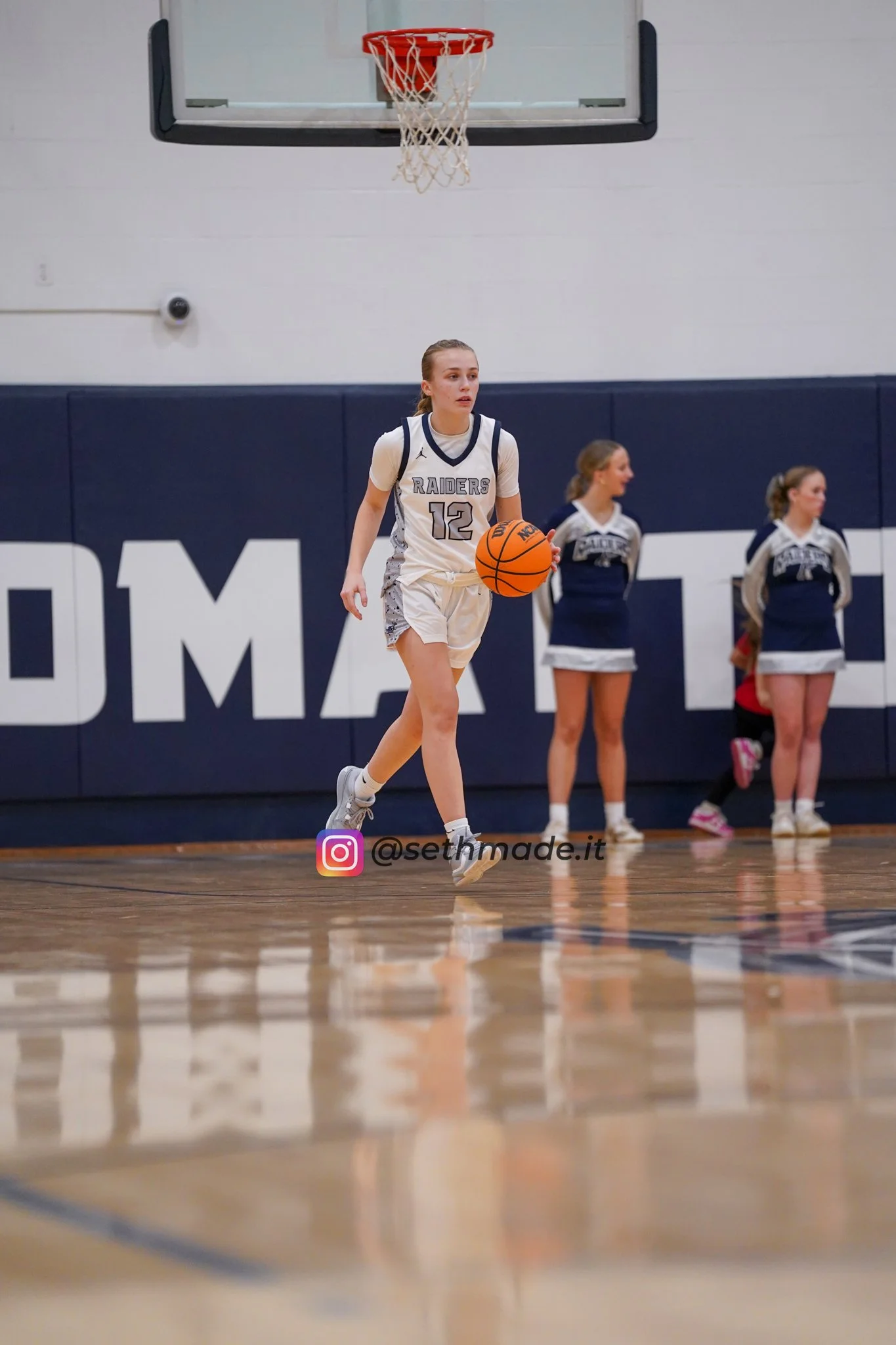 A young female basketball player in a white uniform with the number 12 dribbling a basketball on a court, with cheerleaders standing in the background.