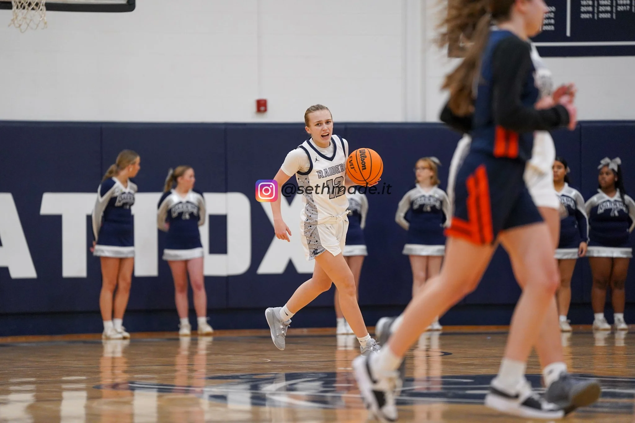 A female basketball player in a white and navy uniform dribbling a basketball on a gym court, with cheerleaders standing in the background against a wall.