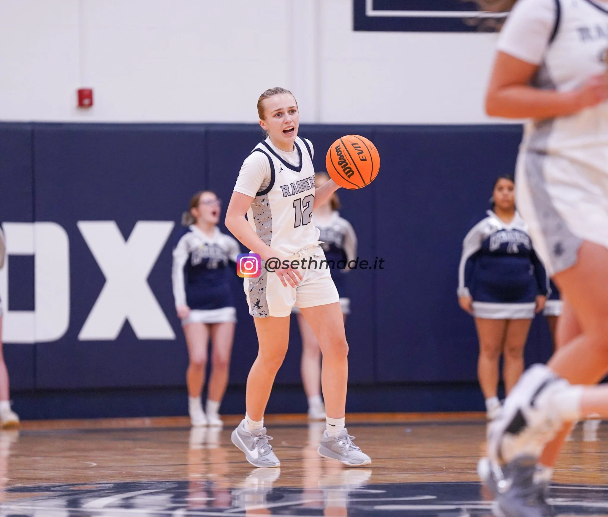 A female basketball player with jersey number 12 in a game, holding a basketball on a court with cheerleaders in the background.