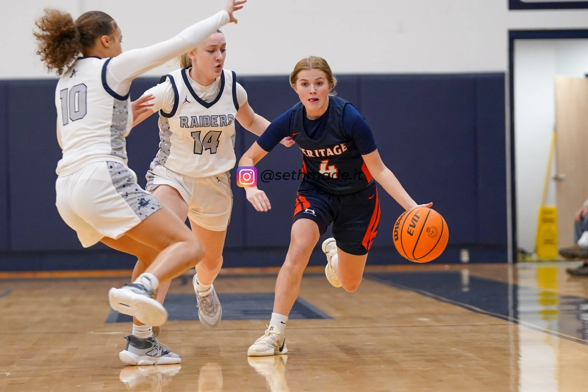 Three young female basketball players are actively engaged in a game on an indoor court. The player in the center, wearing a navy jersey with the number 4 and 'HERITAGE' written on it, is dribbling the basketball and looking forward. Two players in w