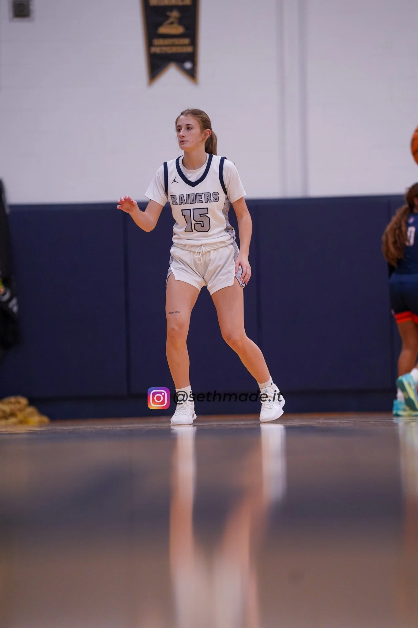A female basketball player with jersey number 15, wearing a White Raiders uniform, standing on a basketball court, mid-game, and looking to her left.