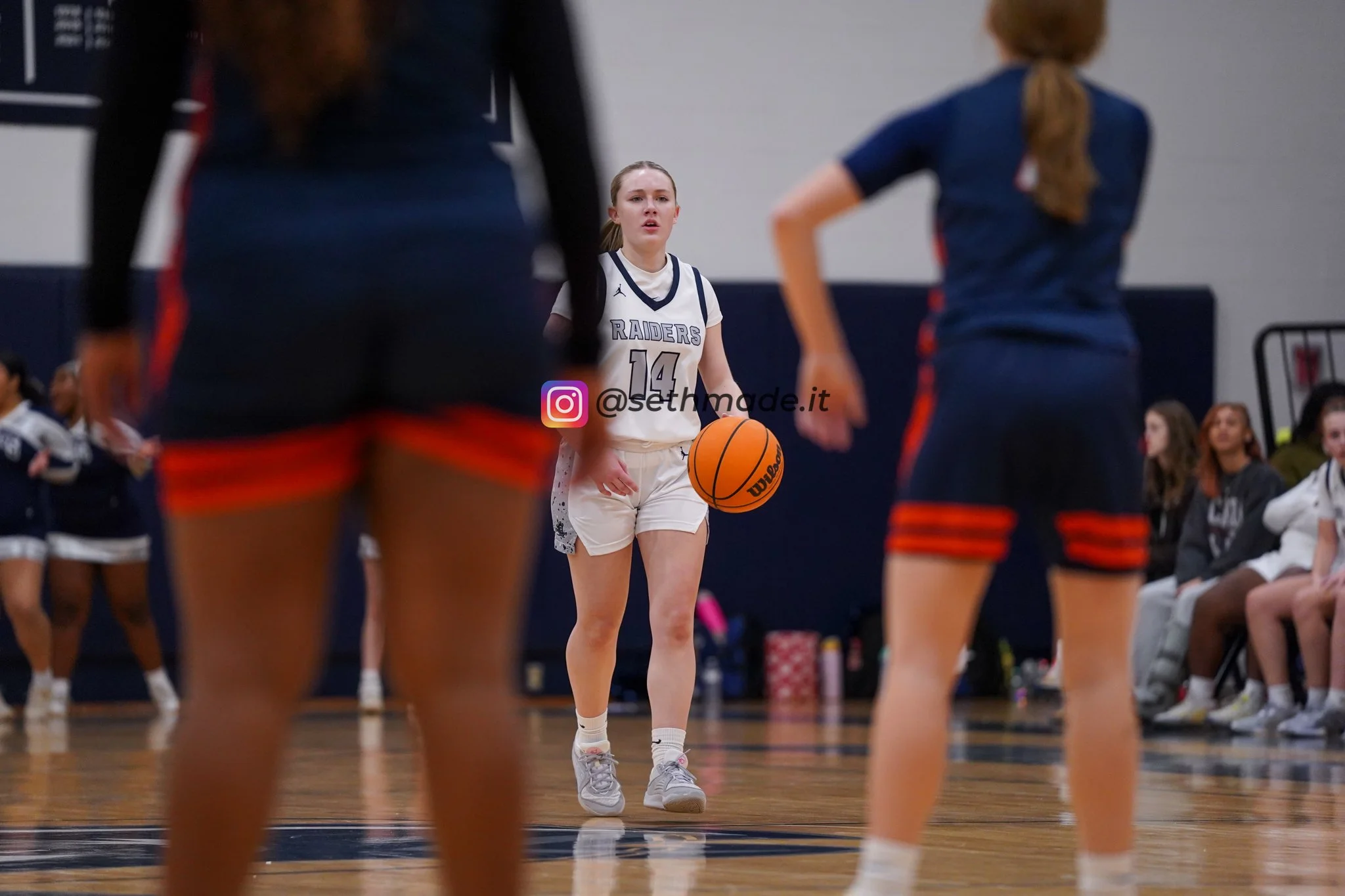 A female basketball player in a white uniform with the number 14, holding a basketball, preparing to make a shot or pass on a gymnasium court. Two other players in dark uniforms are in the foreground, partially blocking the view, with spectators seat