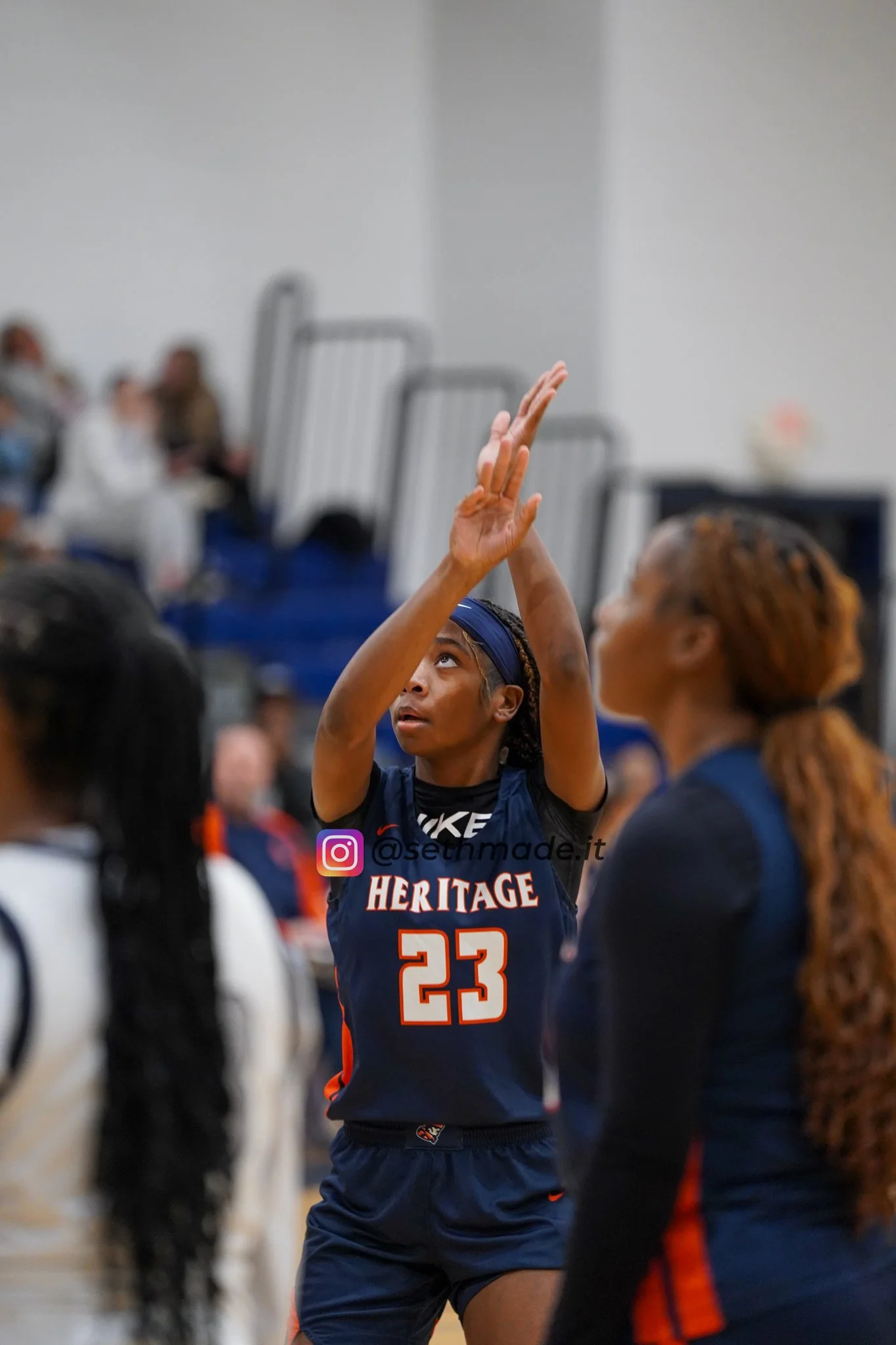 A female basketball player with dark skin, wearing a blue jersey with the number 23, is shooting a basketball during a game. Her hair is tied back with a headband, and she is focused on her shot. Other players and spectators are visible in the backgr