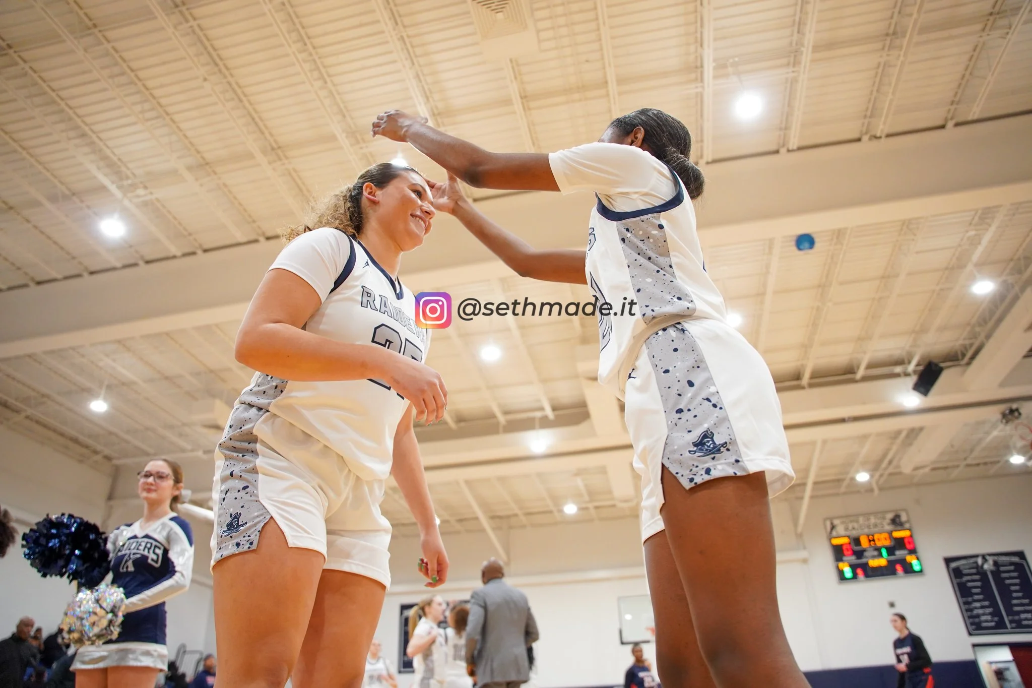 Two female basketball players wearing white jerseys are celebrating together on the court, one is touching the other's head while smiling, in a gymnasium with people and a scoreboard in the background.