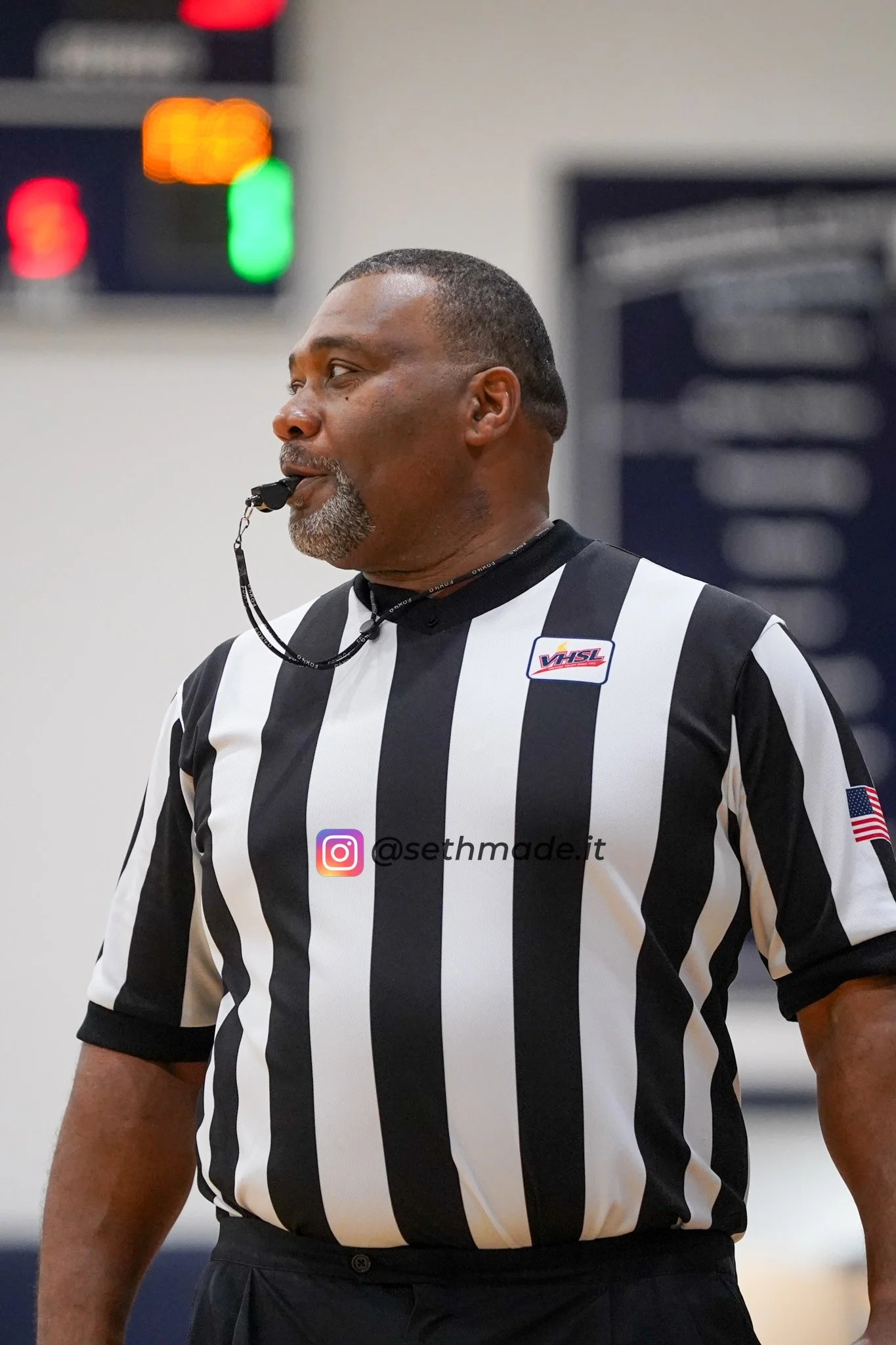 A referee wearing a black and white striped jersey with the VHSL logo, the American flag on the sleeve, and an Instagram handle on the front, officiating at a sports event.