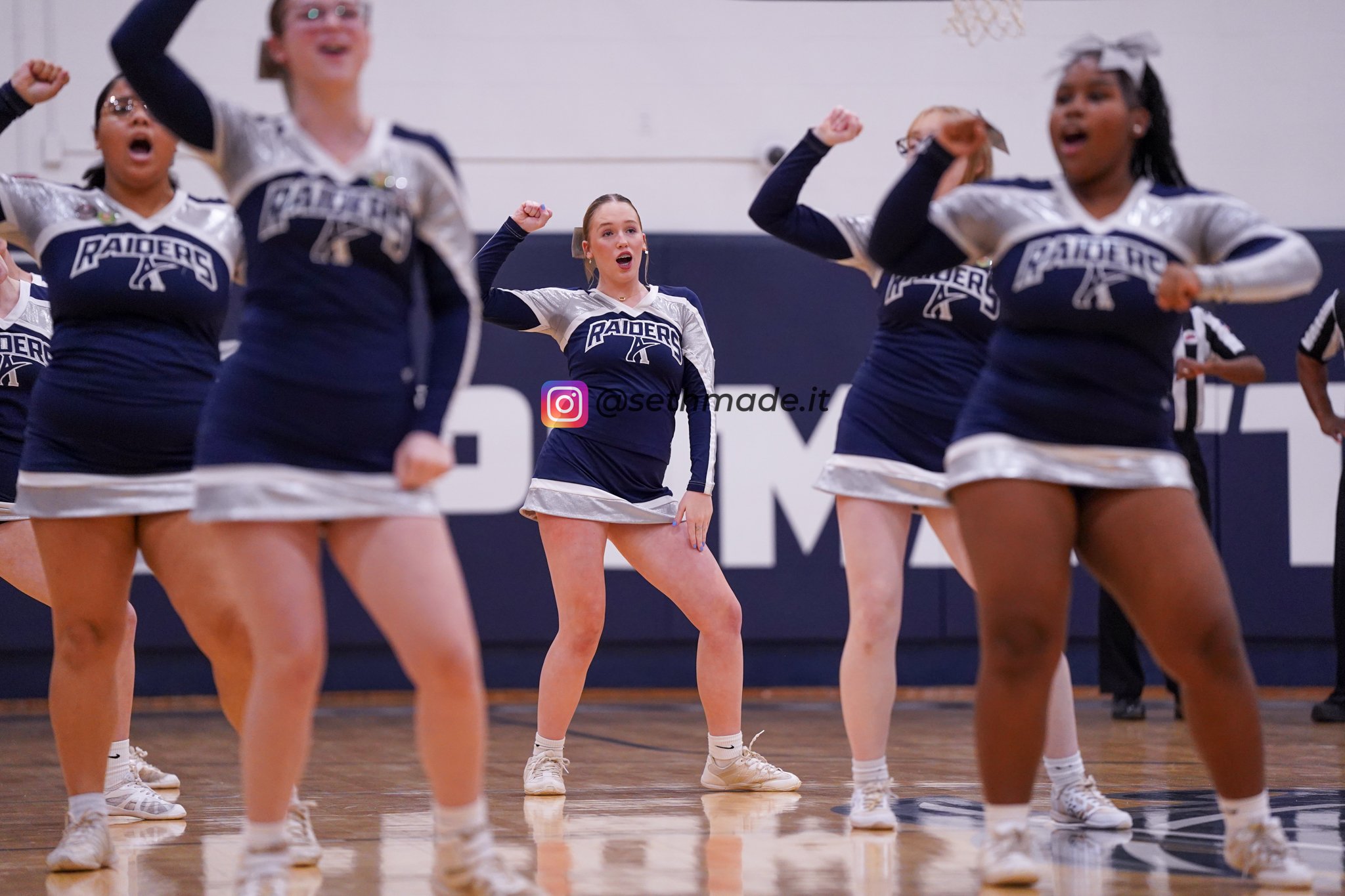 Cheerleaders performing a routine in a gymnasium, wearing blue and white uniforms with 'Raiders' on the front, with their arms flexed and excited expressions.