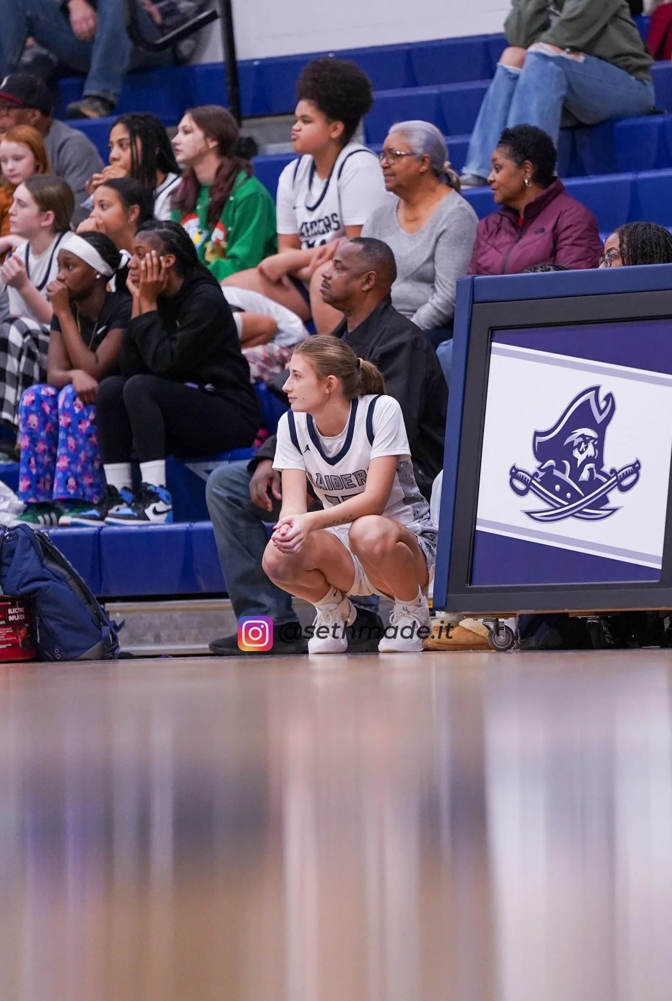 A young female basketball player in a white and navy uniform squats on the sidelines of a basketball game, watching the game intently while seated near a monitor with a logo of a pirate and crossed swords. Behind her, a diverse group of spectators, i