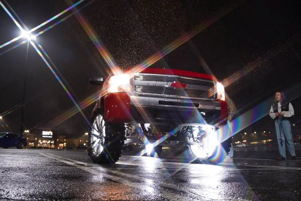 A red Chevrolet truck with bright headlights on in a parking lot at night, with a woman standing nearby.