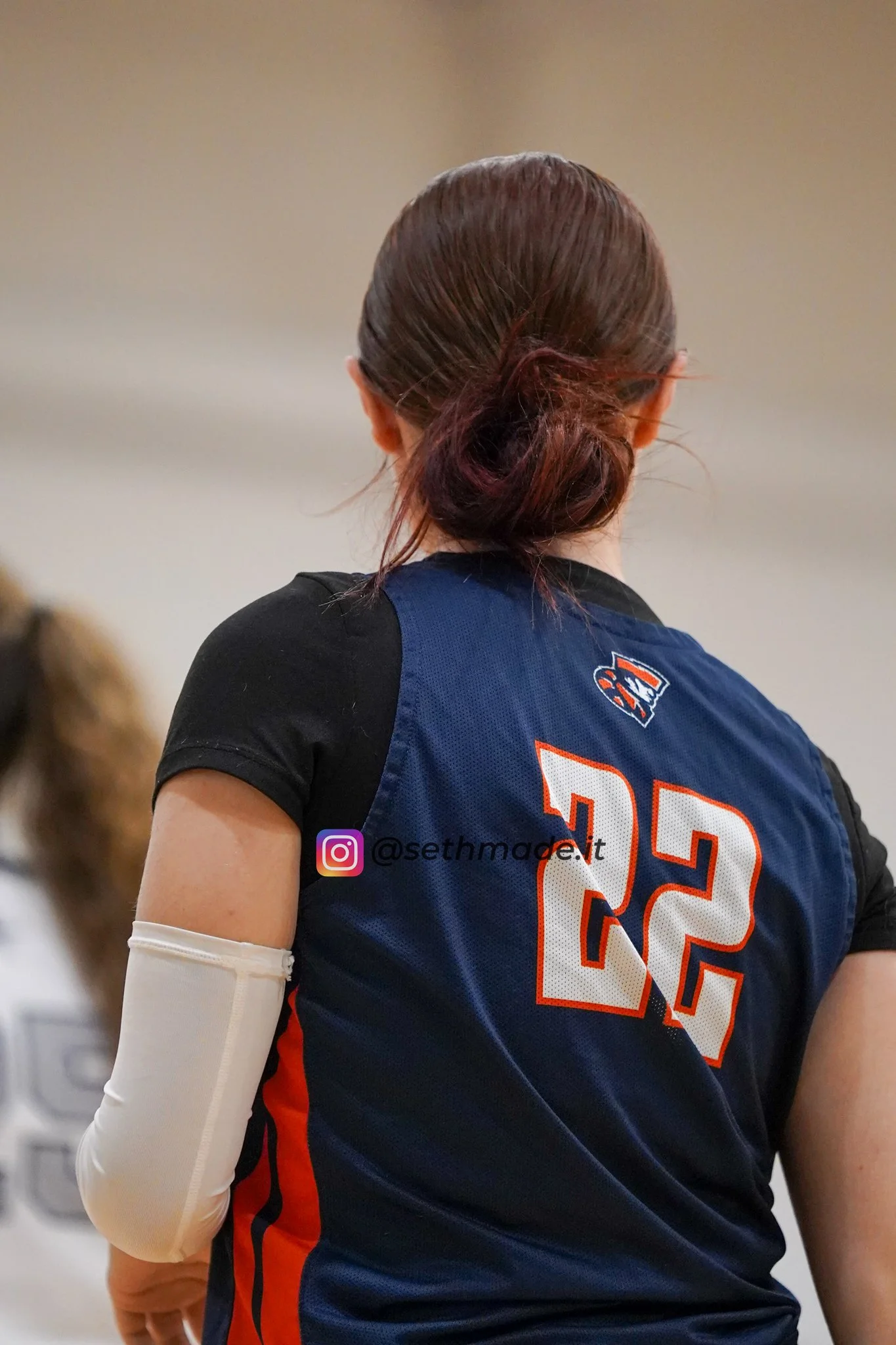 Back of female volleyball player wearing a navy blue and black uniform with the number 22 on it, with long brown hair tied in a ponytail, and a white arm sleeve.
