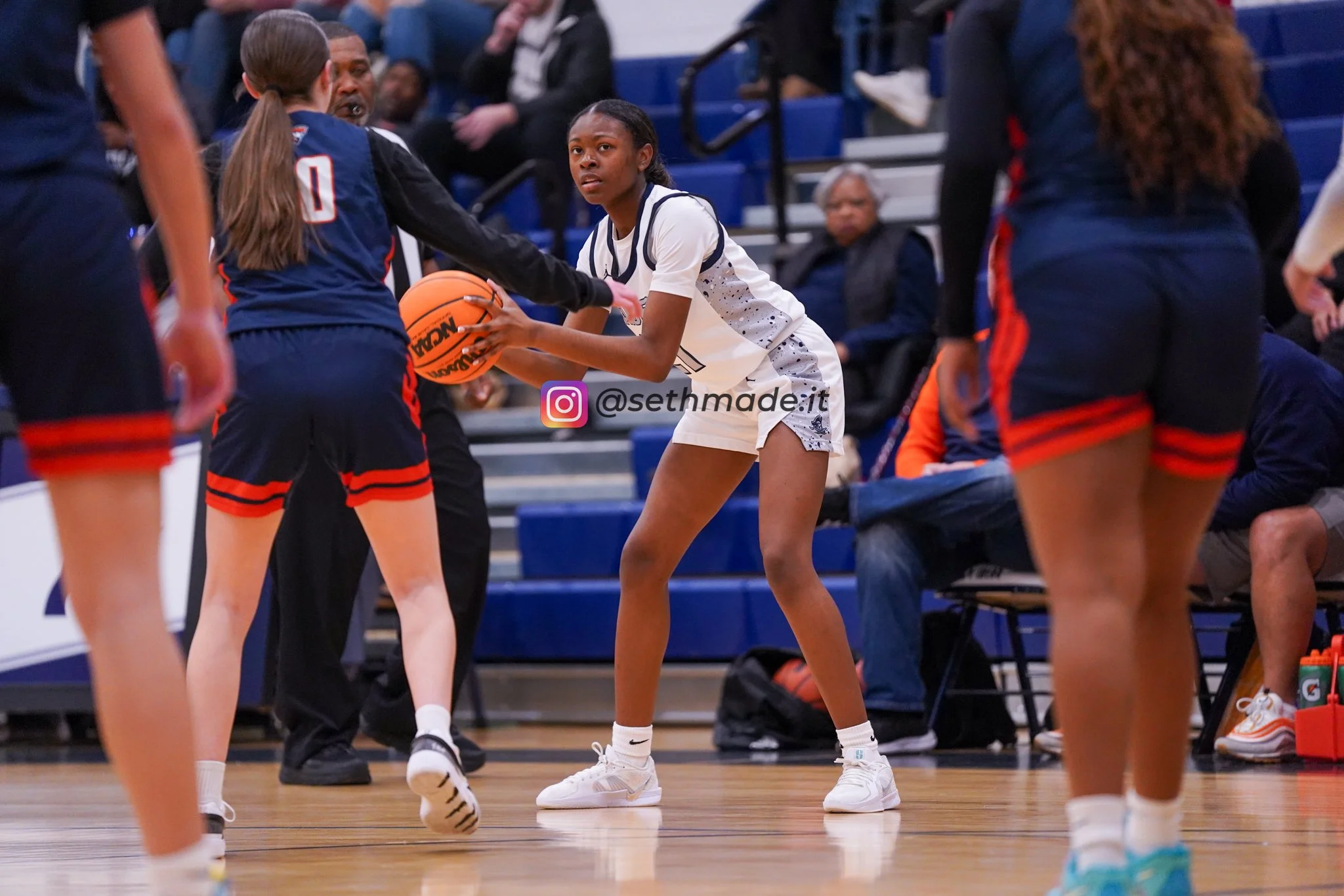 A girl in a white basketball uniform holding a basketball, guarded by other players during a basketball game.
