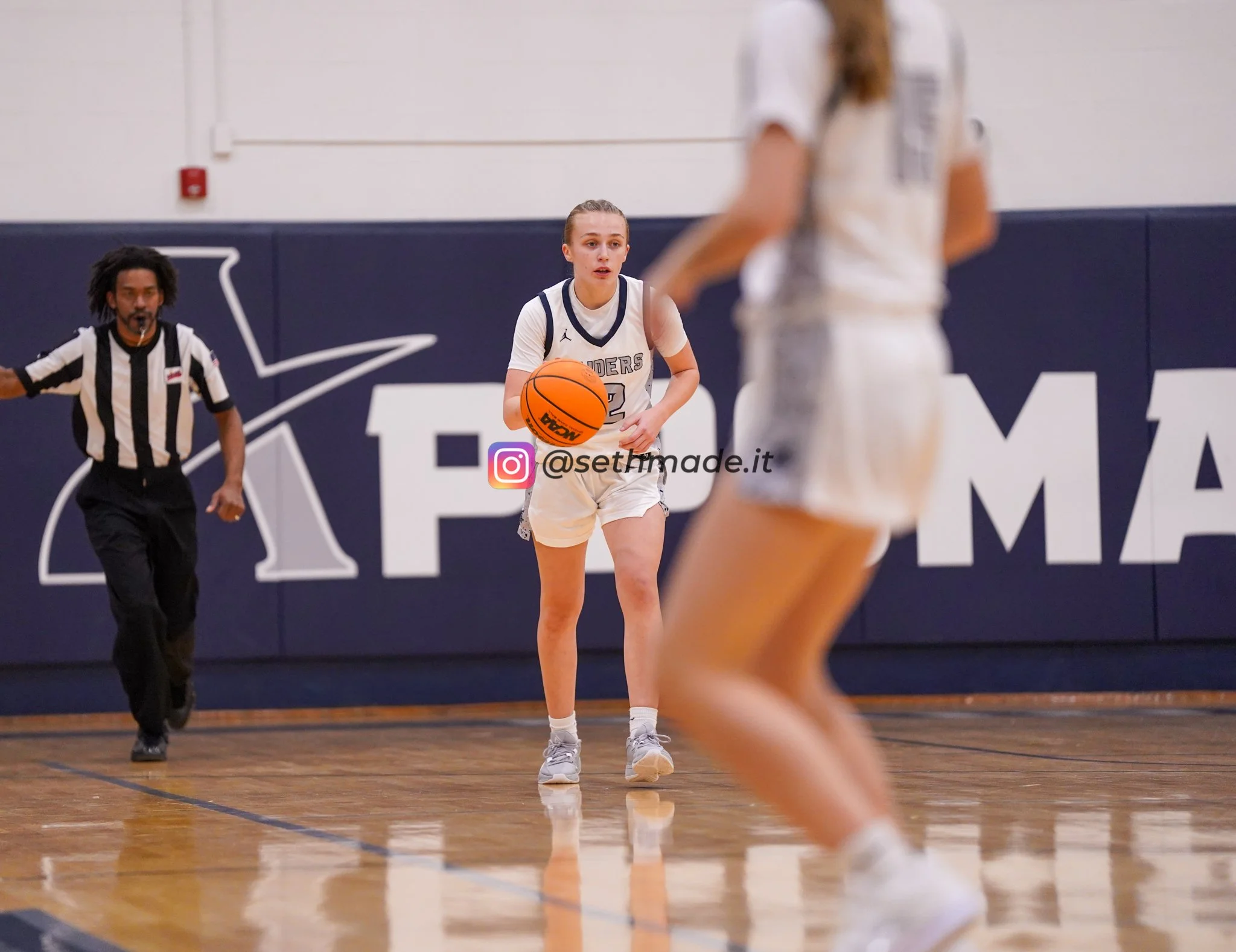 A female basketball player in a white uniform dribbling the ball during a game, with a female teammate partially visible in the foreground and a referee running behind her, on a wooden court with a blue wall and logo in the background.