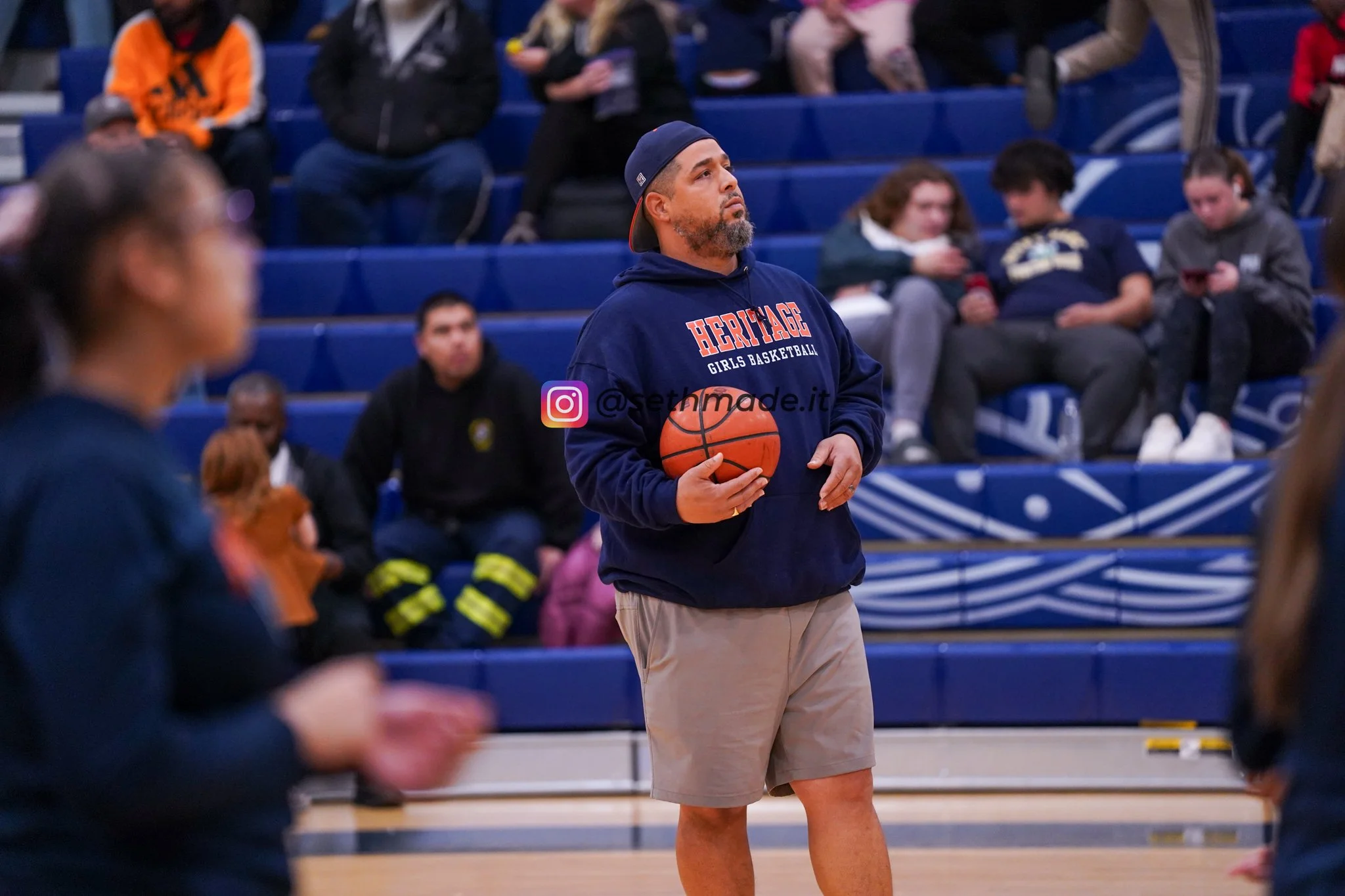 A man in a navy blue hoodie and shorts holding a basketball, standing in a gymnasium during a basketball practice or game, with spectators in the background.