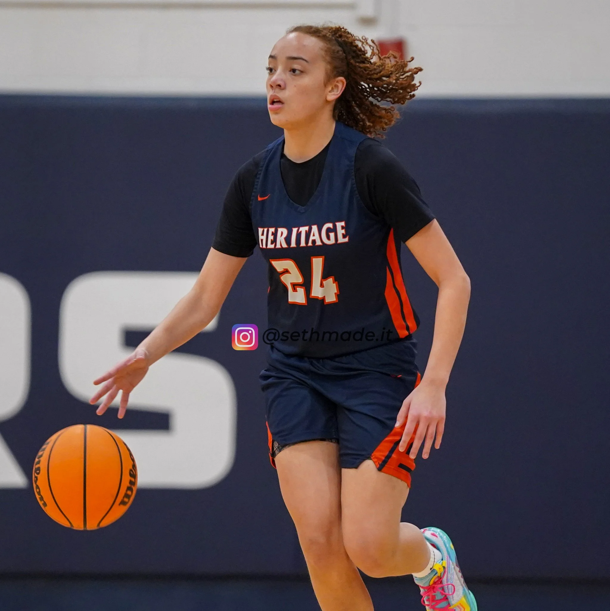 Young female basketball player in navy and orange jersey dribbling a basketball on an indoor court.