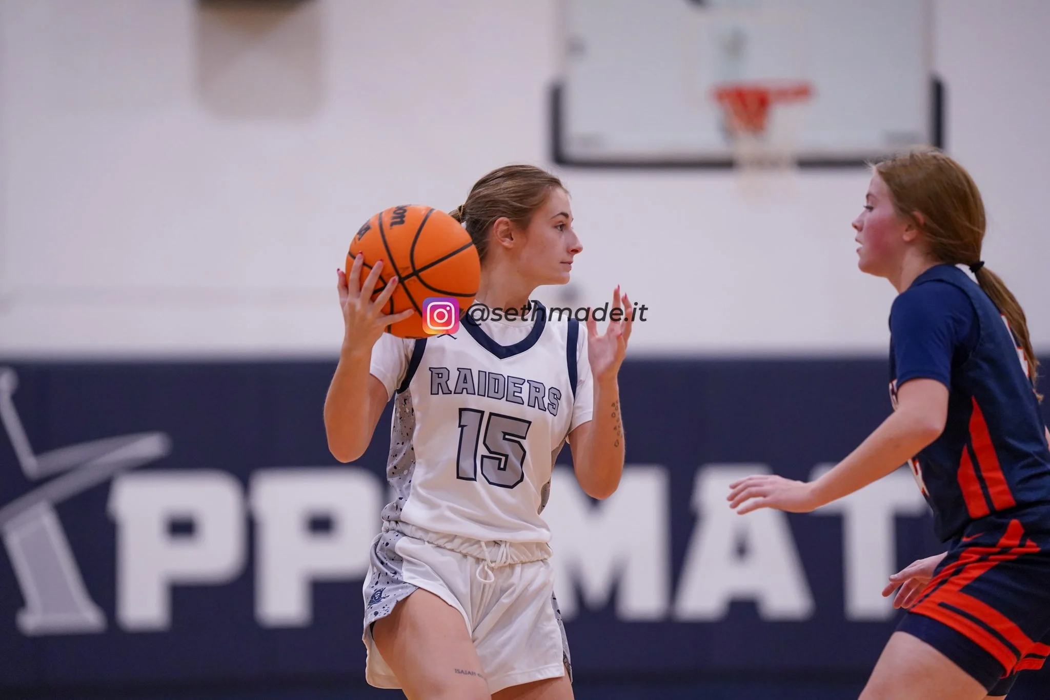 A female basketball player in a white and purple uniform with the number 15, holding a basketball near her shoulder, faces another female player in a navy blue uniform with red accents on a basketball court.