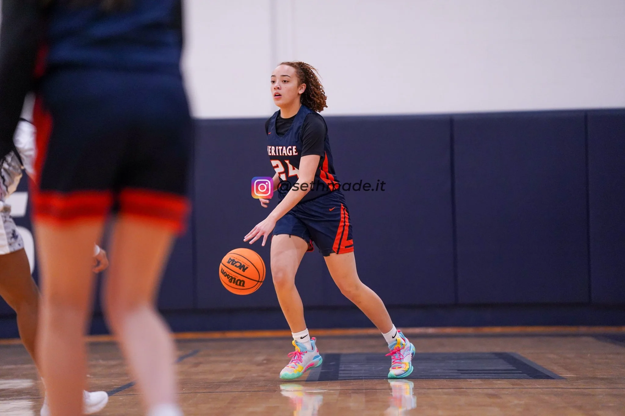 A young woman in a navy and orange basketball uniform dribbling a basketball on an indoor court, with other players partially visible nearby.