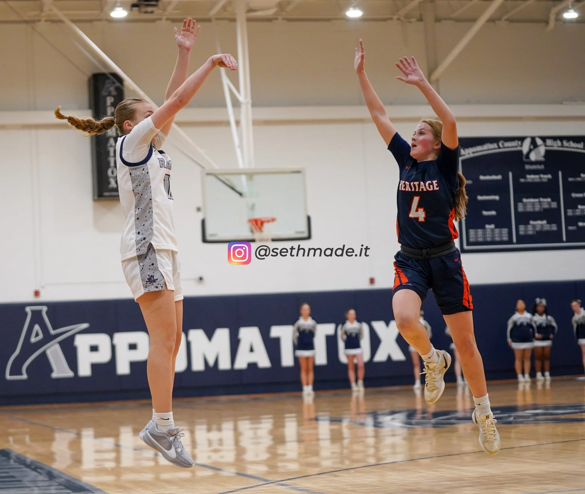 Two female volleyball players are jumping and reaching for the ball during a game in an indoor gymnasium. One player is in a white jersey with the number 0, and the other is in a navy blue jersey with the number 4. Cheerleaders line up along the wall