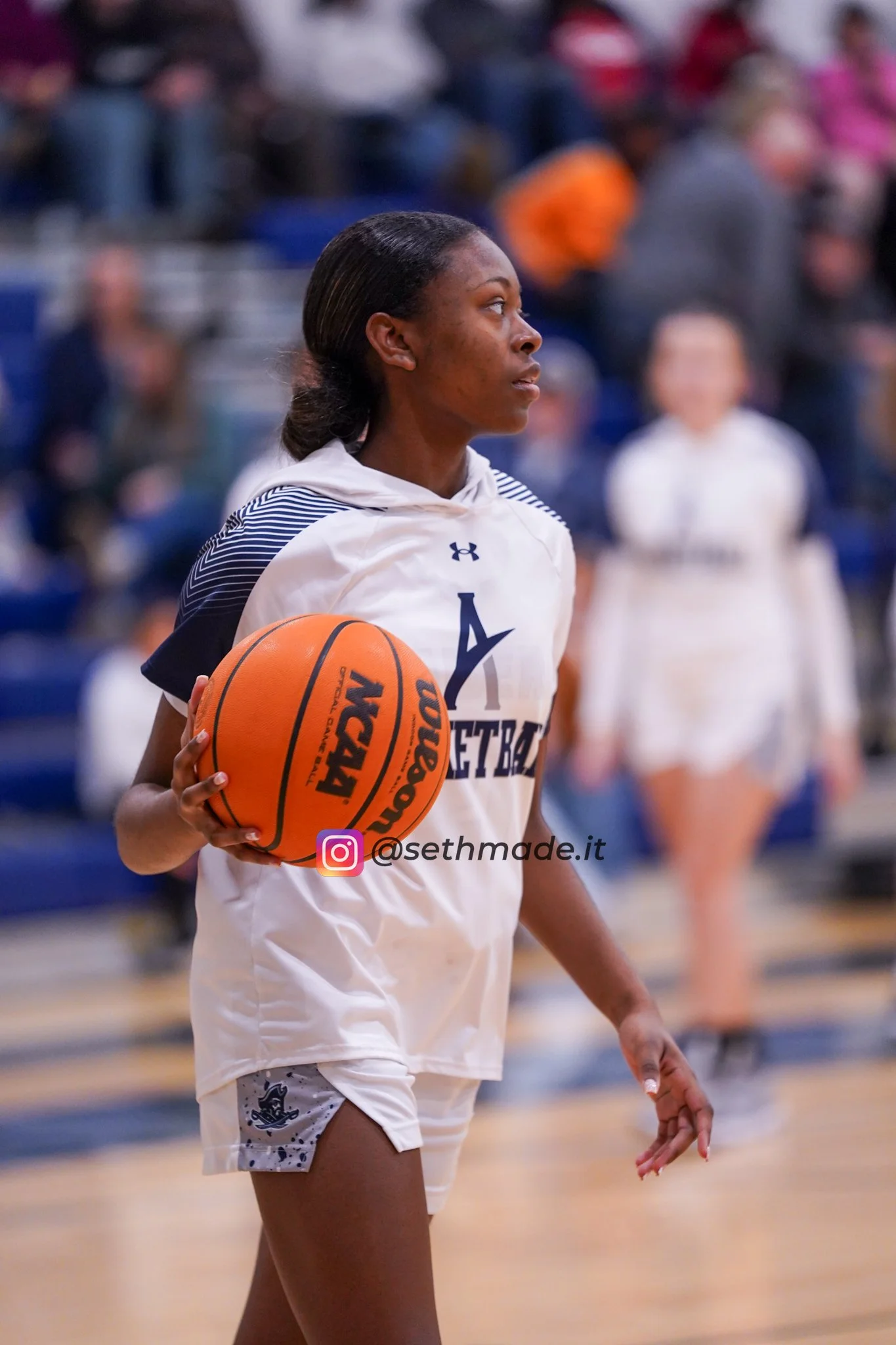 A female basketball player in a white uniform with the words 'A BASKETBALL' on it, holding an orange basketball, standing on an indoor court with blurred spectators in the background.