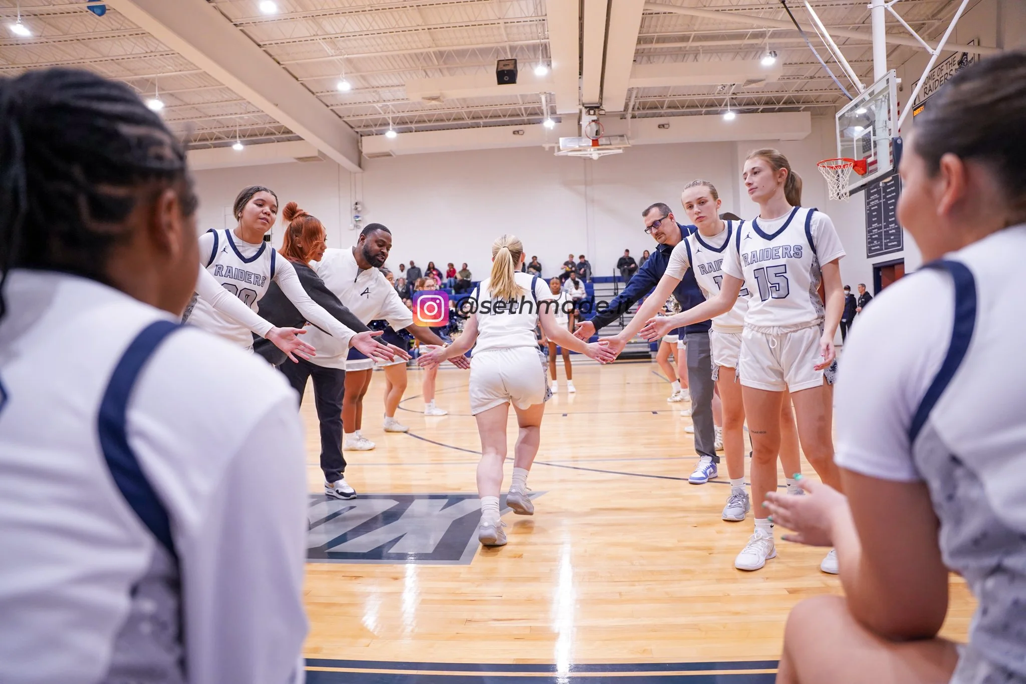 A women's basketball team on the court during a timeout, lined up in a tunnel for a cheer or motivational moment, with players and coaches reaching out to each other.