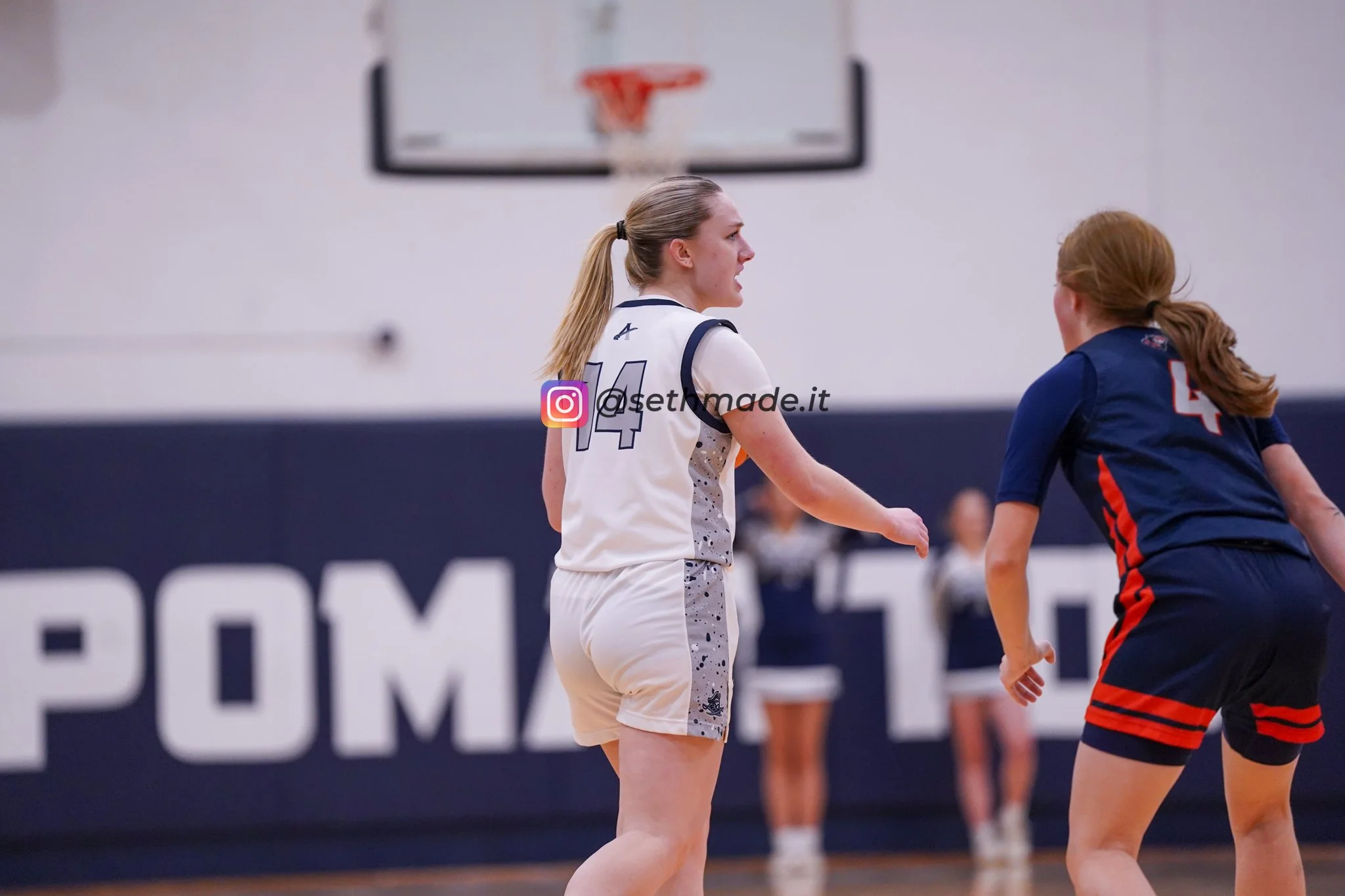 A female volleyball player with blonde hair in a ponytail, wearing a white jersey with the number 14, is shouting during a game. She is facing another player in a dark blue jersey with the number 4. The scene takes place in an indoor gymnasium with a