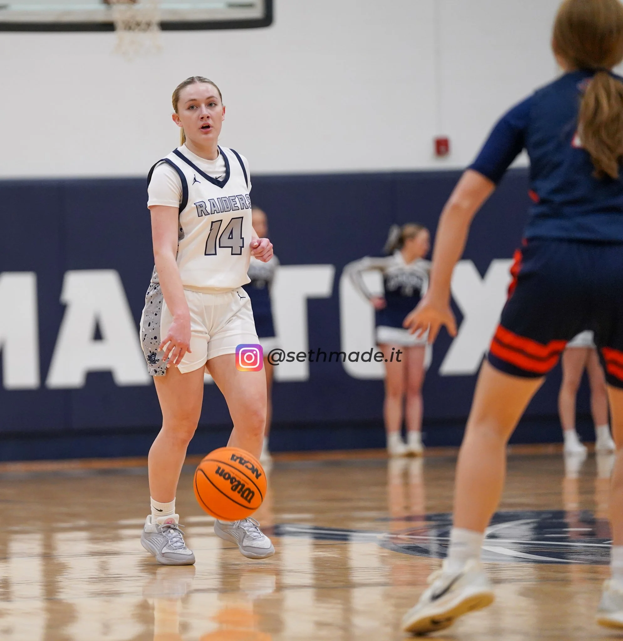 A female basketball player wearing a white and gray uniform with the number 14 dribbling an orange basketball on a gym court while facing other players.