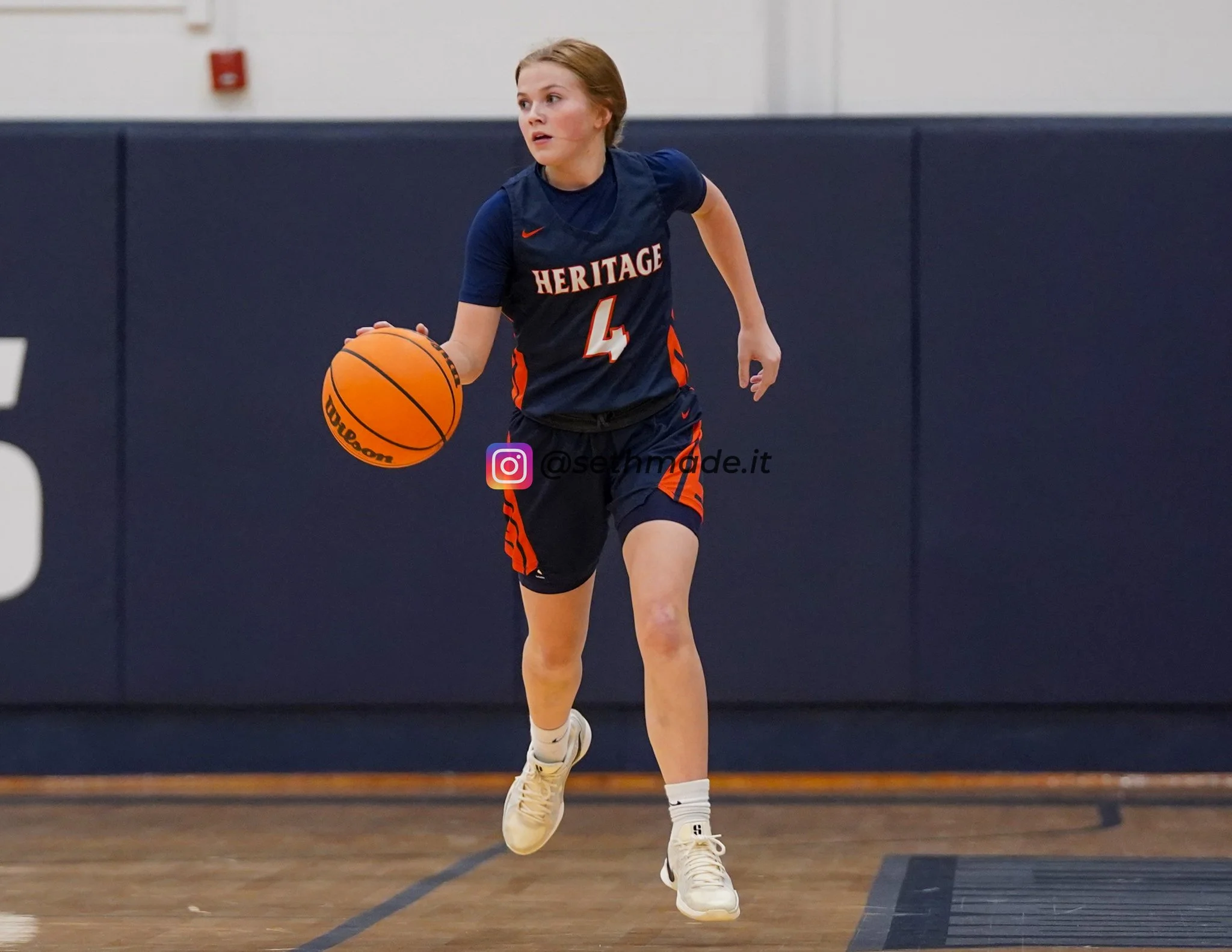 A female basketball player in a navy blue and orange jersey dribbling a basketball on an indoor court.