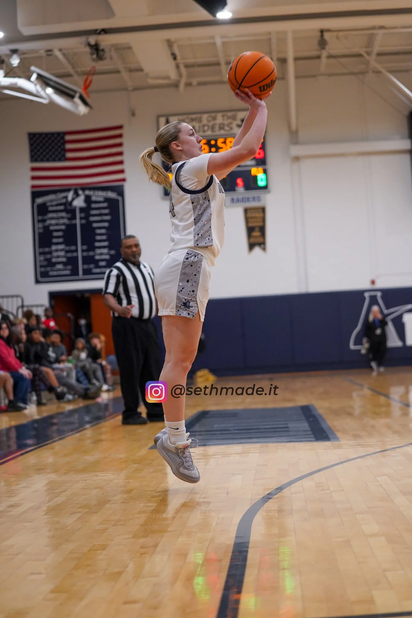 A female basketball player in a white and gray uniform is jumping to shoot the basketball during a game in a gymnasium.