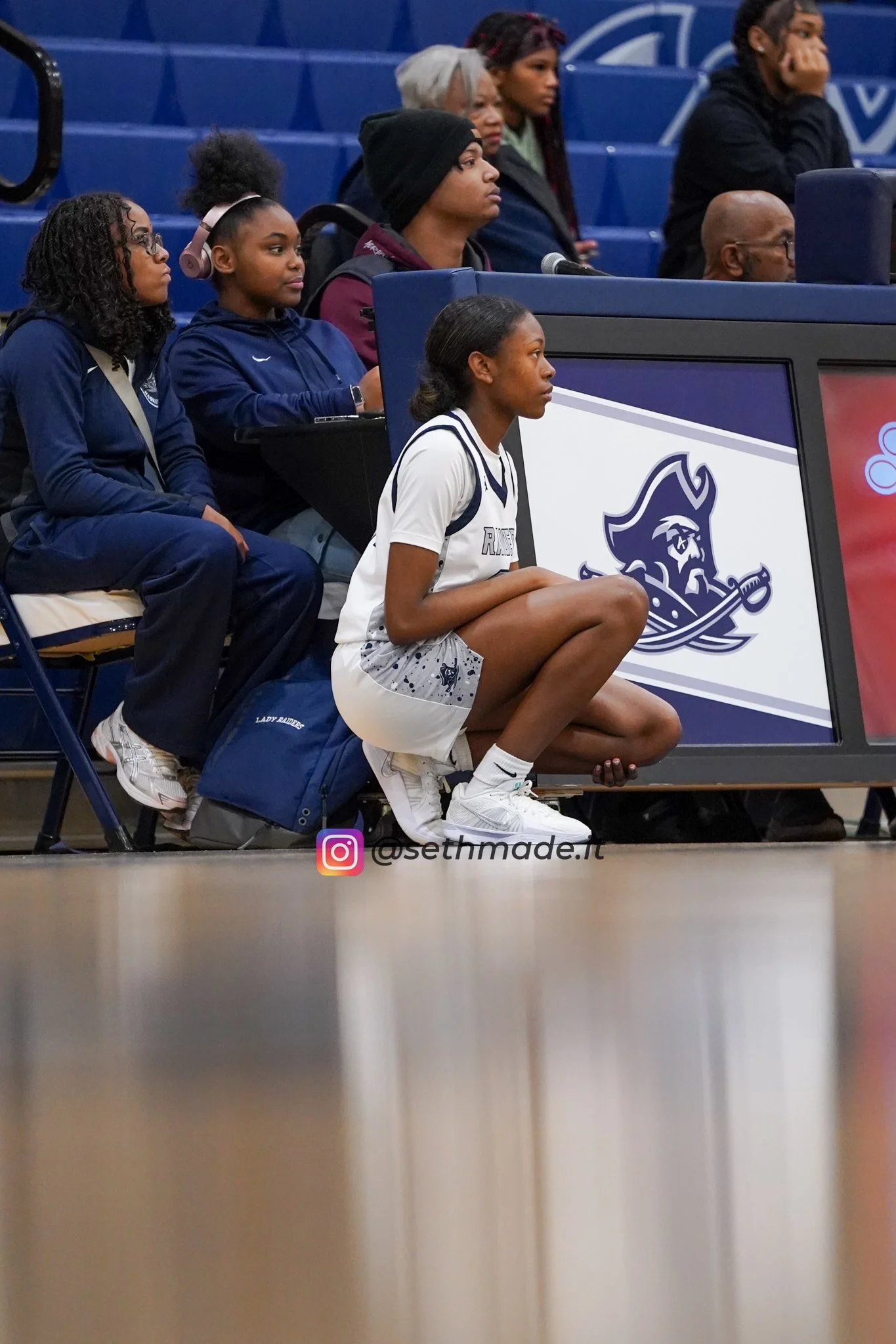 A group of young women, likely athletes, sitting on the benches and watching a game in a gymnasium. One girl in a white jersey is crouching on the floor near a digital scoreboard, which displays a logo of a pirate with a sword.