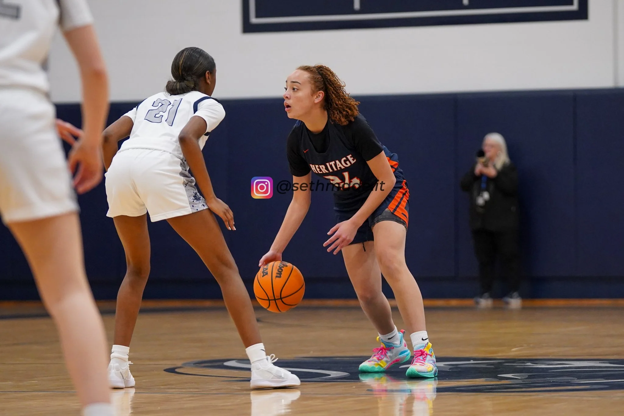 A female basketball player with curly hair and brightly colored sneakers dribbling the basketball, while a young opponent in a white uniform defends her on the court.