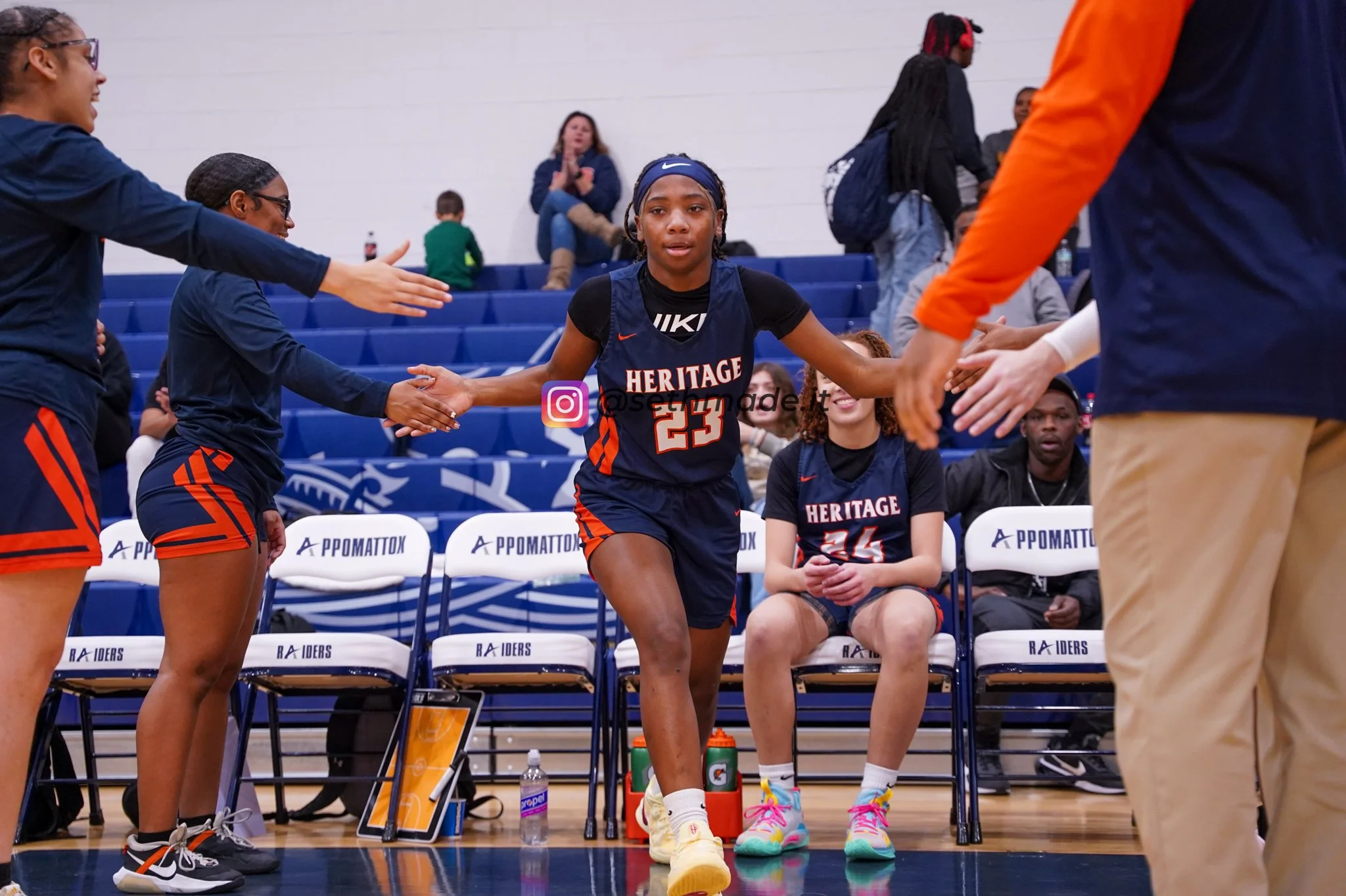 A female basketball player with jersey number 23 high-fives her teammates during a game or practice, seated on a bench in a gymnasium.