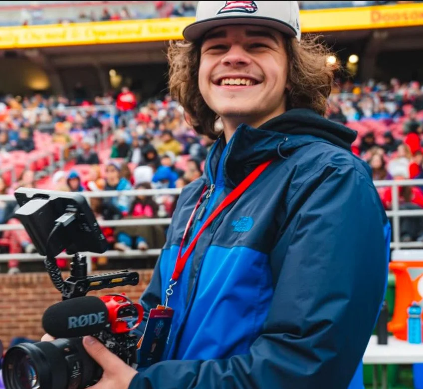 A young man with curly hair and a cap smiling at a sporting event, holding a camera with a microphone labeled RODE and an external monitor, with a crowd in the background.