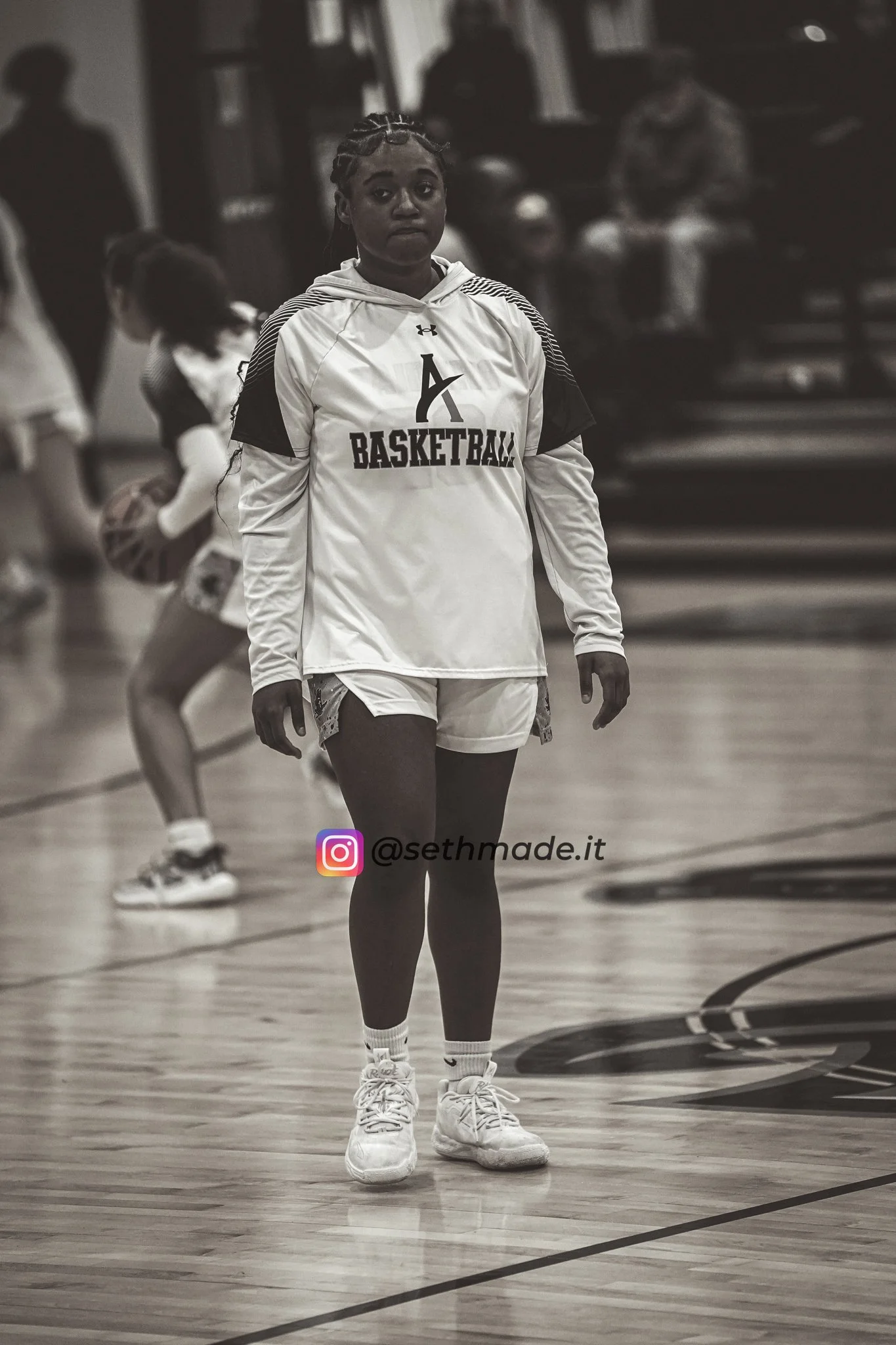 Black and white photo of a young female basketball player standing on an indoor court in athletic wear, with other players practicing in the background.