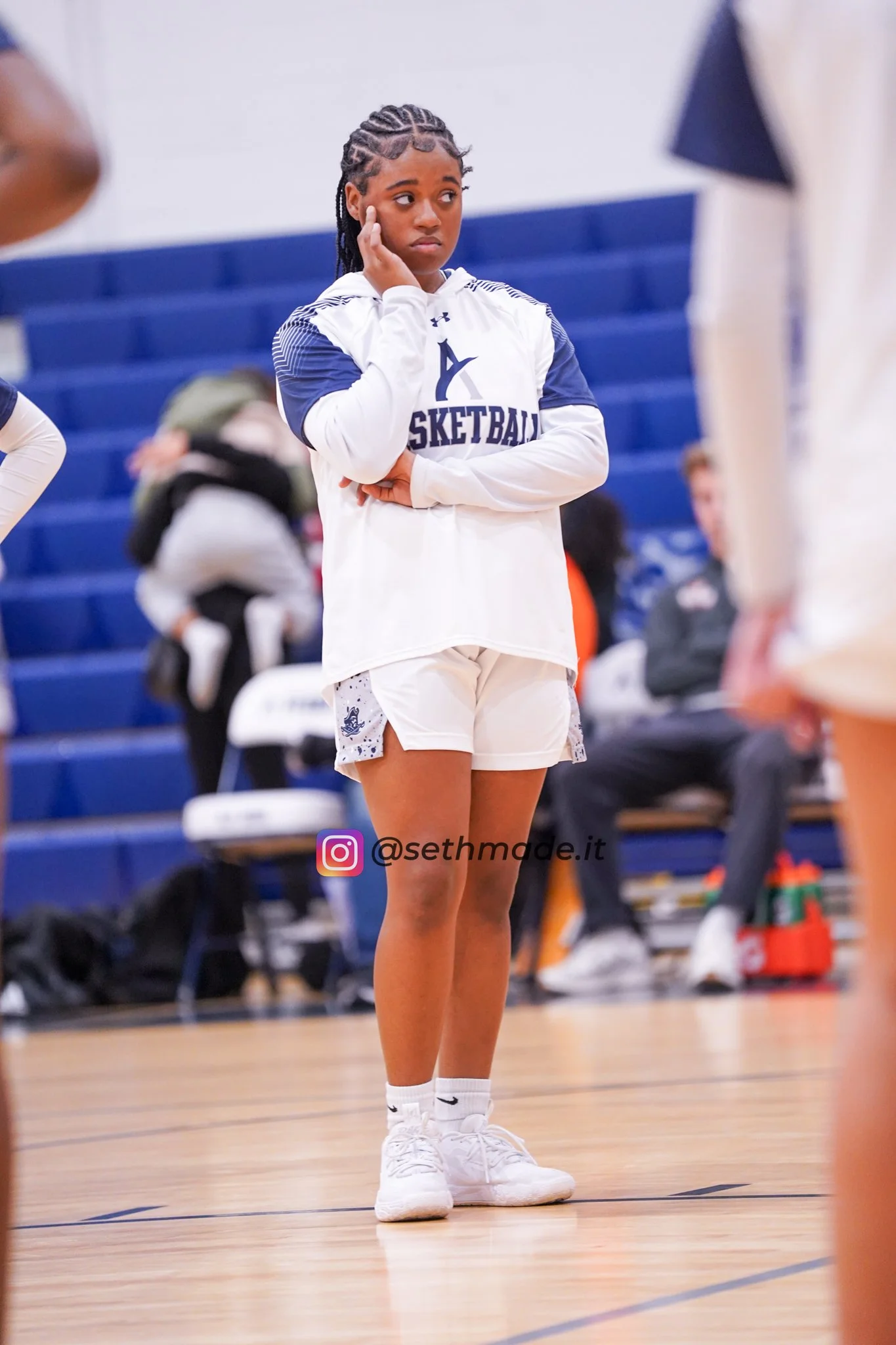 A young girl standing on a basketball court, wearing a white and navy blue basketball uniform, with her hand on her face, appearing thoughtful or concerned.