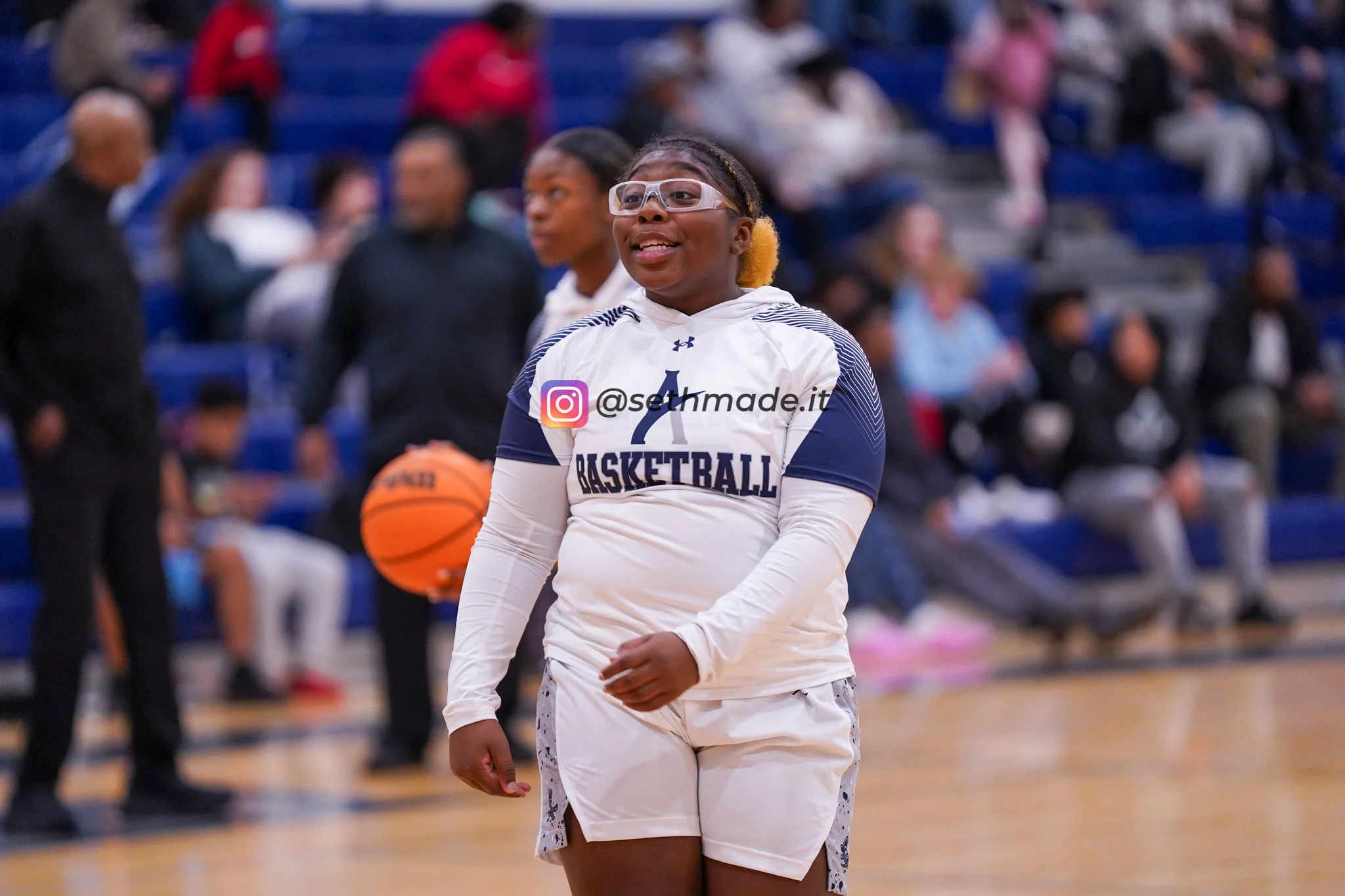 A young woman wearing glasses, a white and navy basketball jersey, and white shorts stands on a basketball court, with spectators in the background.