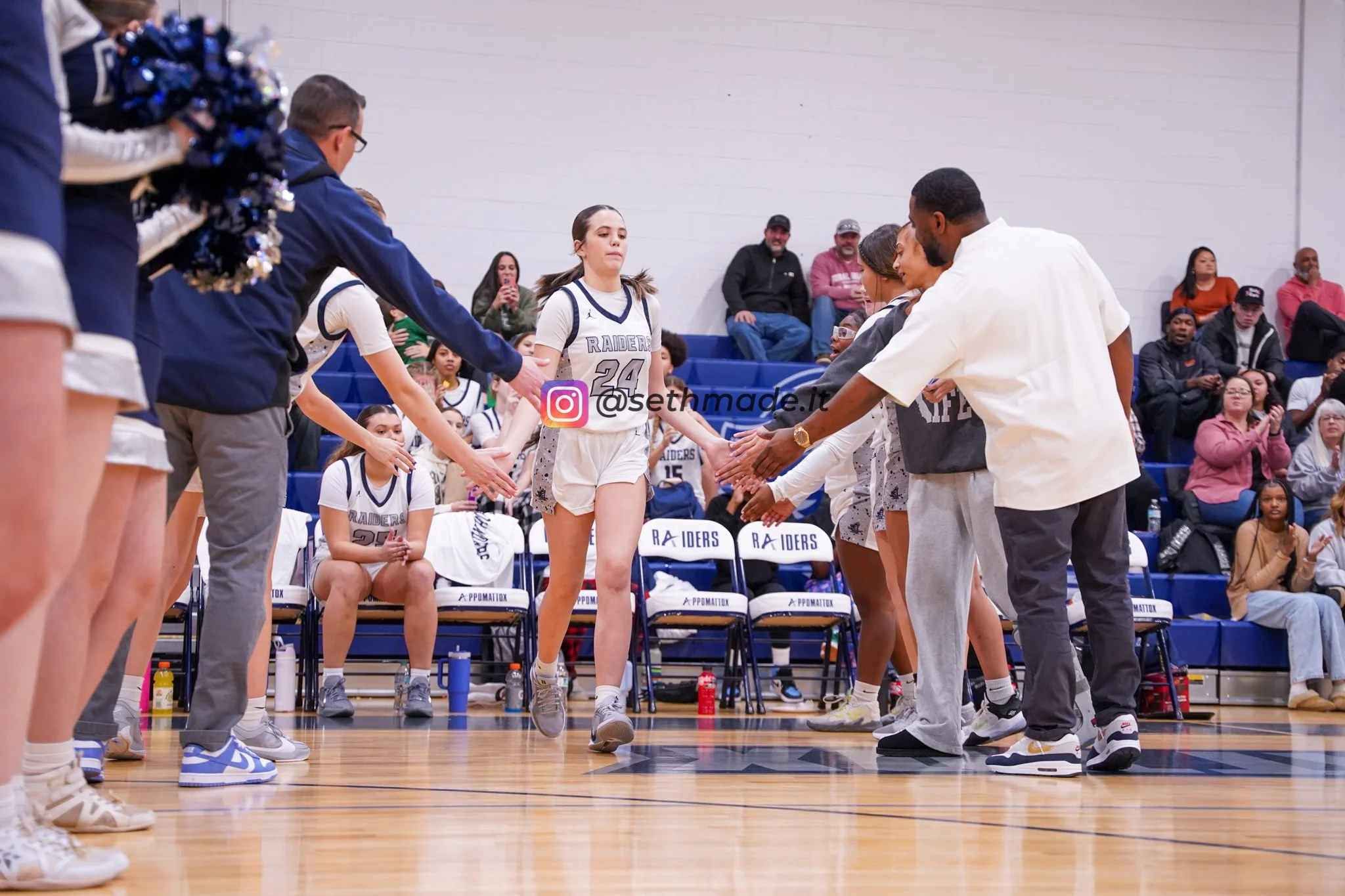 A basketball player in a white uniform with the number 24, standing on a court and preparing for a game or event, surrounded by coaches and teammates, with spectators sitting in the background.