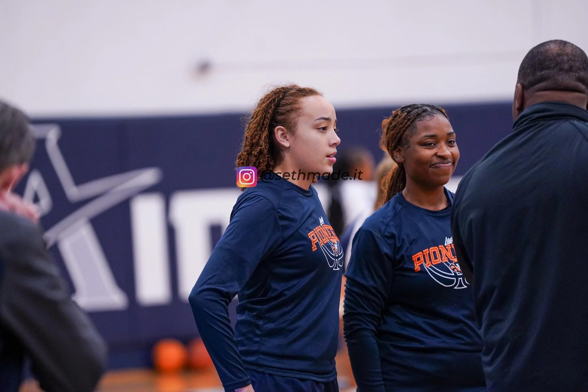 Two female basketball players in navy blue team uniforms are listening to a coach during a timeout in an indoor gymnasium. One player has red hair and the other has brown braided hair.