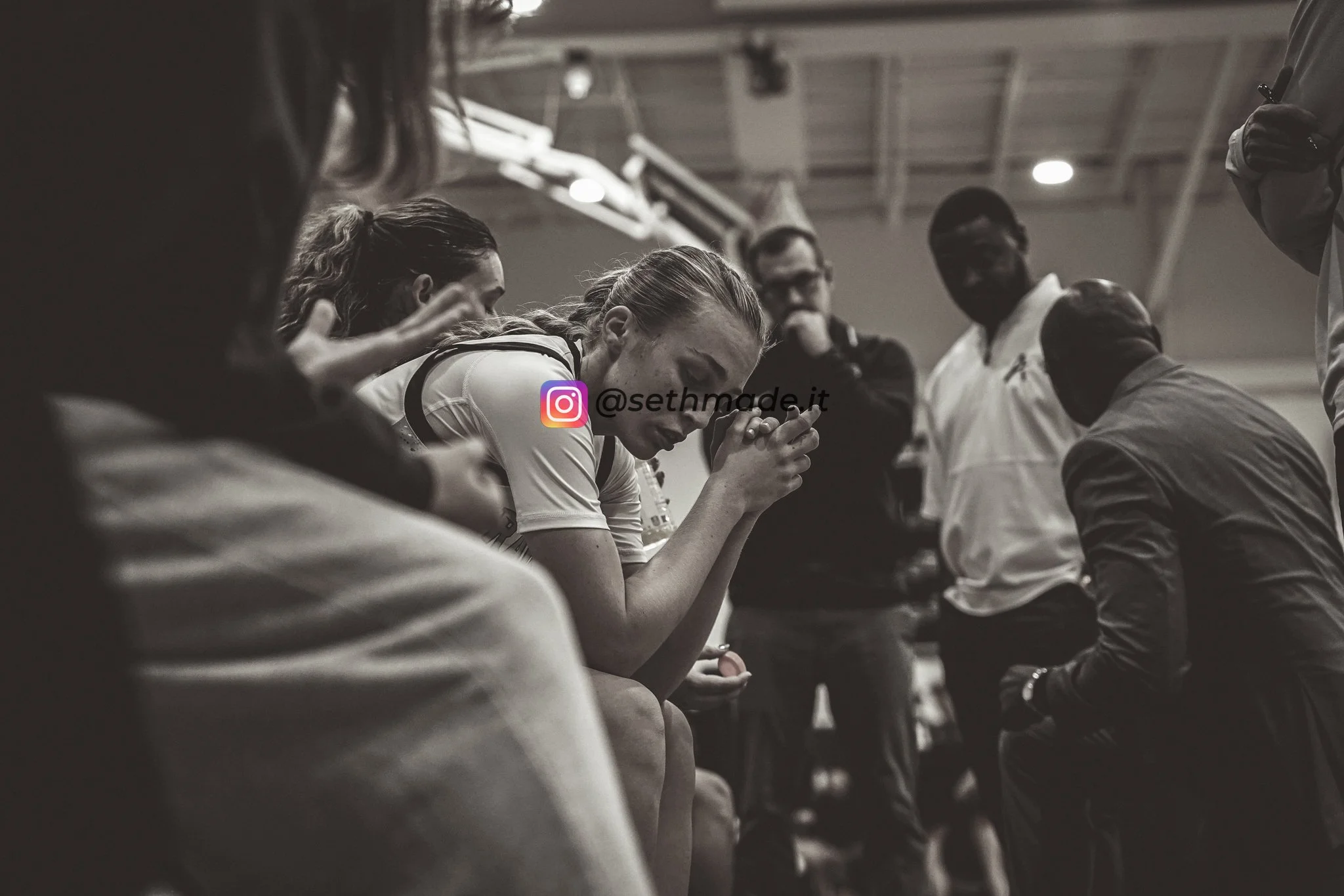 Black and white photo of a woman in a sports uniform with her head bowed and hands clasped in prayer, sitting on a bench surrounded by teammates and coaches in a gymnasium.