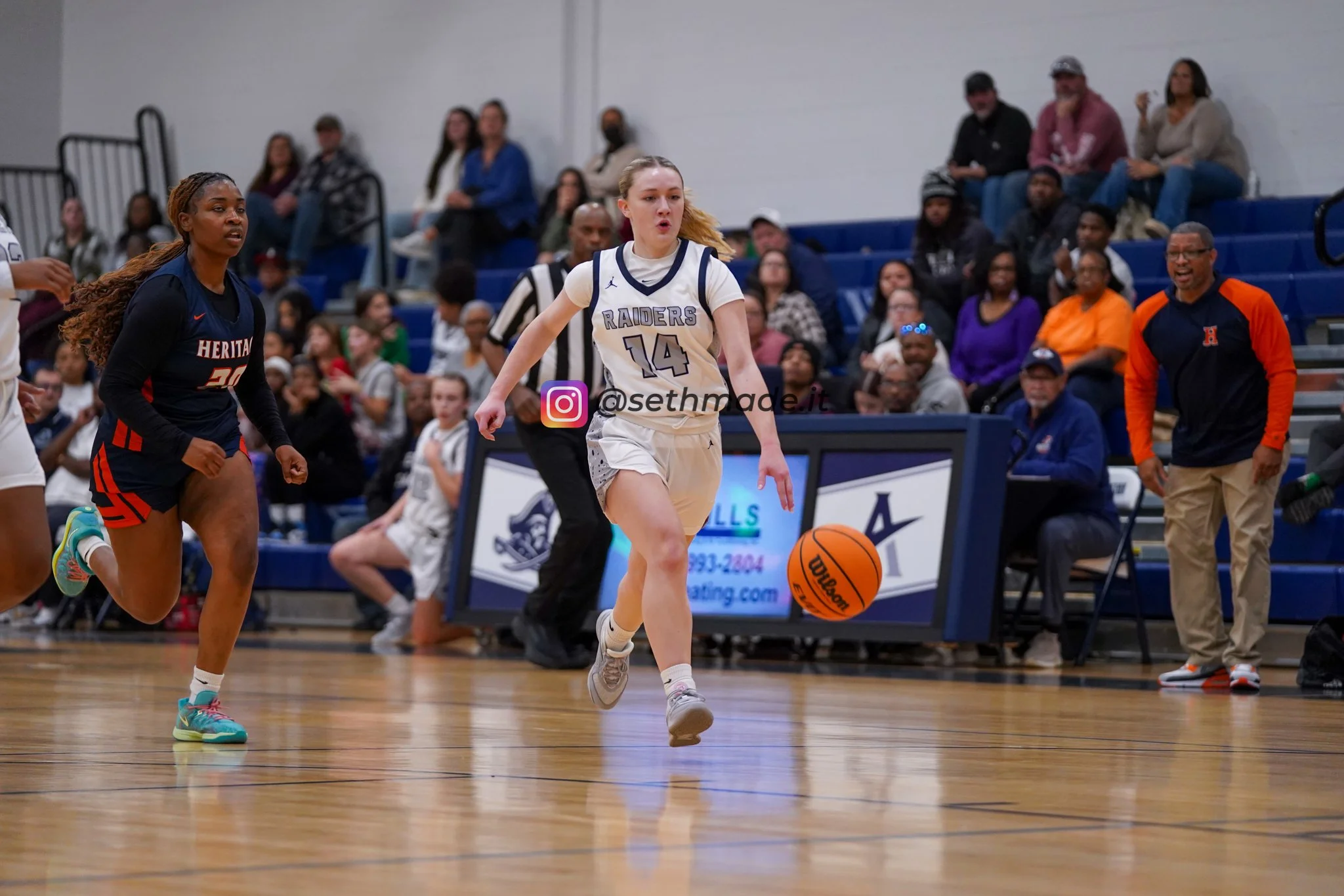 A female basketball player with jersey number 14 dribbling the ball on the court during a game, with fans and team staff watching from the bleachers.
