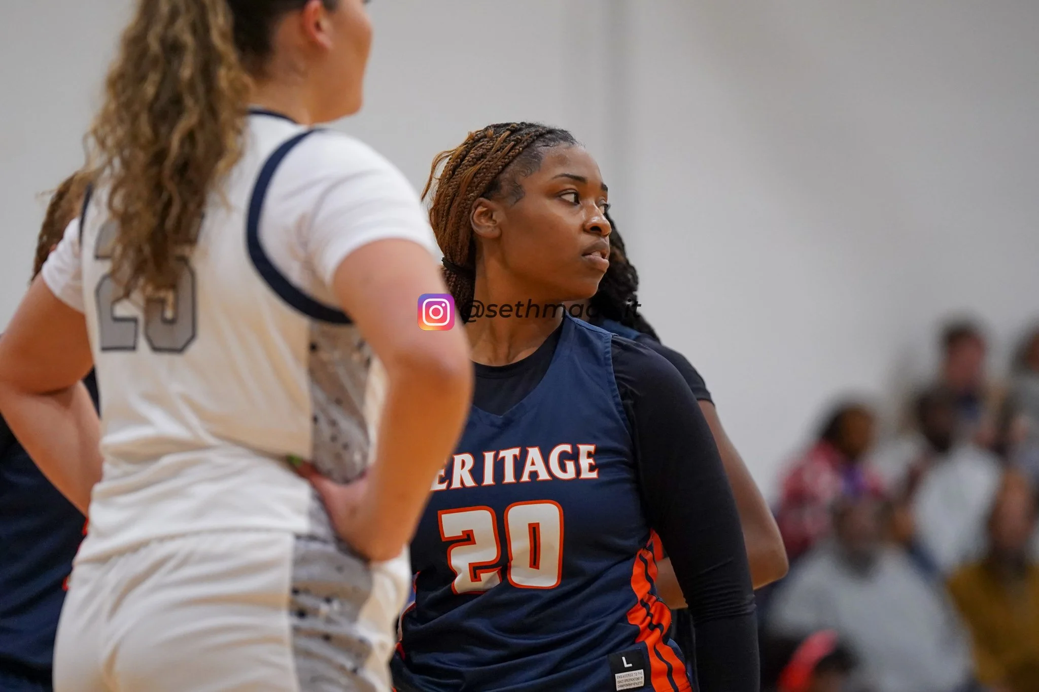 A female basketball player in a blue jersey with the number 20 and the word 'HERITAGE' on it, appears to be in a game, standing on the court with her side to the camera. In the foreground, part of another player in a white jersey with the number 23 i