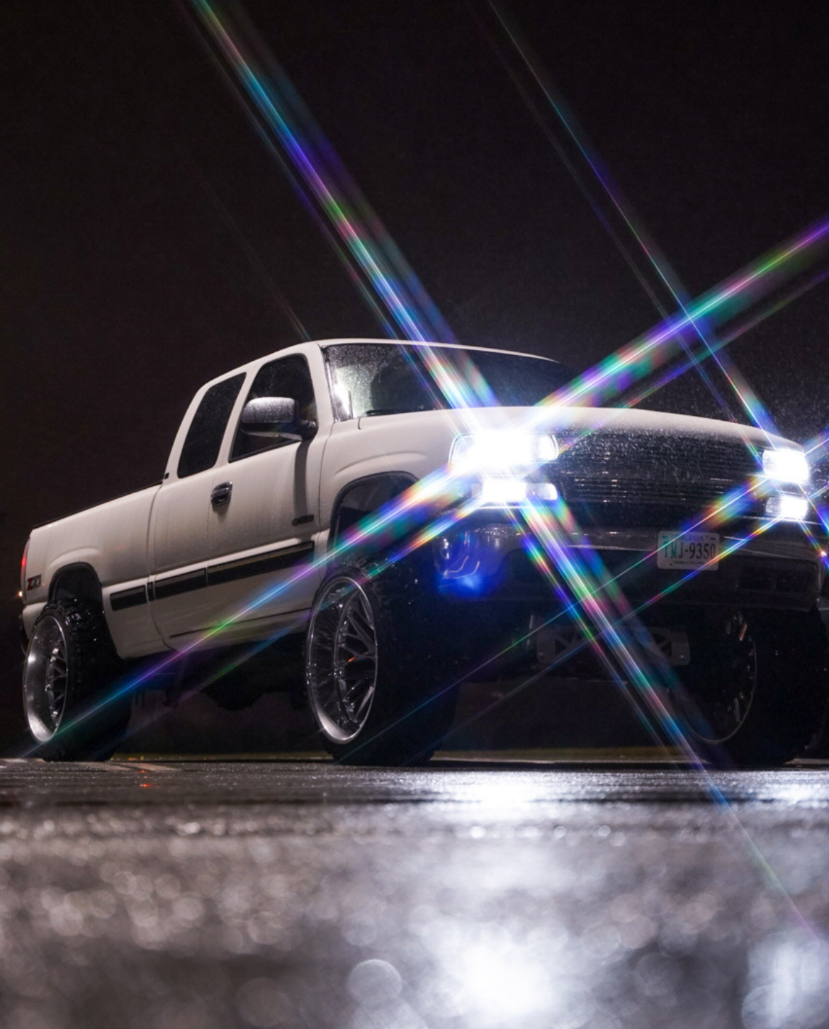 A white pickup truck with shiny wheels at night, with bright headlights and light flares creating rainbow-colored streaks across the scene.