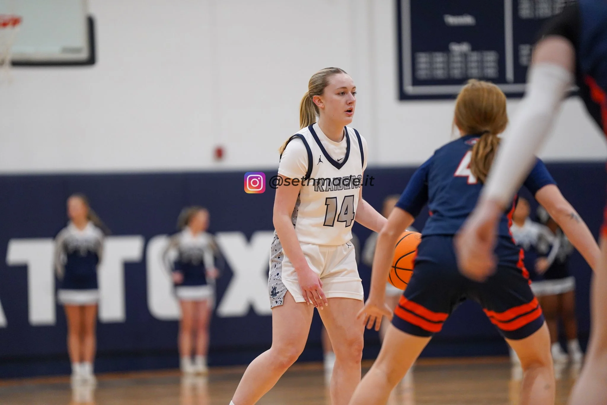 A female basketball player wearing a white jersey with the number 14 is on a court during a game, facing an opponent in a blue and orange uniform, with cheerleaders in the background.