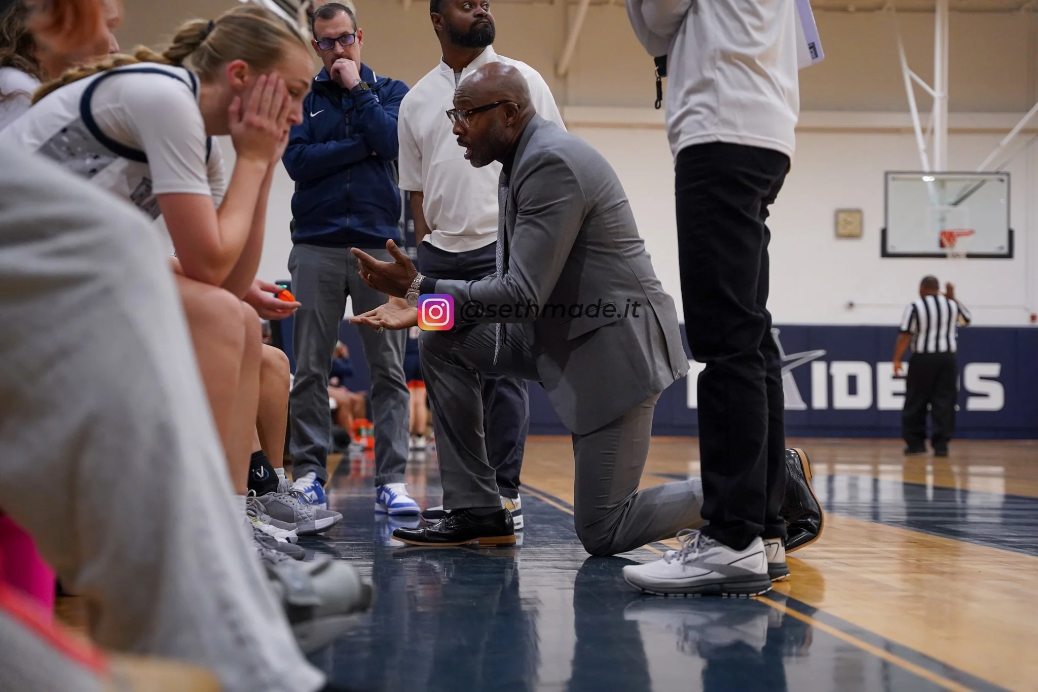 Basketball coach coaching a team during a timeout, kneeling on the court and speaking to players sitting on the bench, with several staff members standing and paying attention, inside a gymnasium.