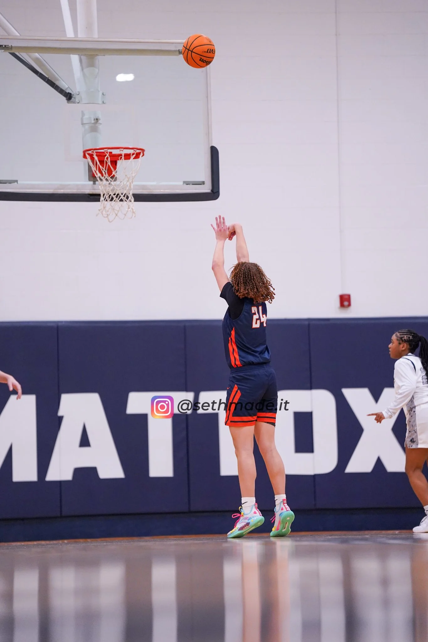 A girl in a navy and orange basketball uniform taking a shot at the hoop in an indoor gym, with the basketball mid-air about to go into the hoop. There is a young girl on the right side watching.