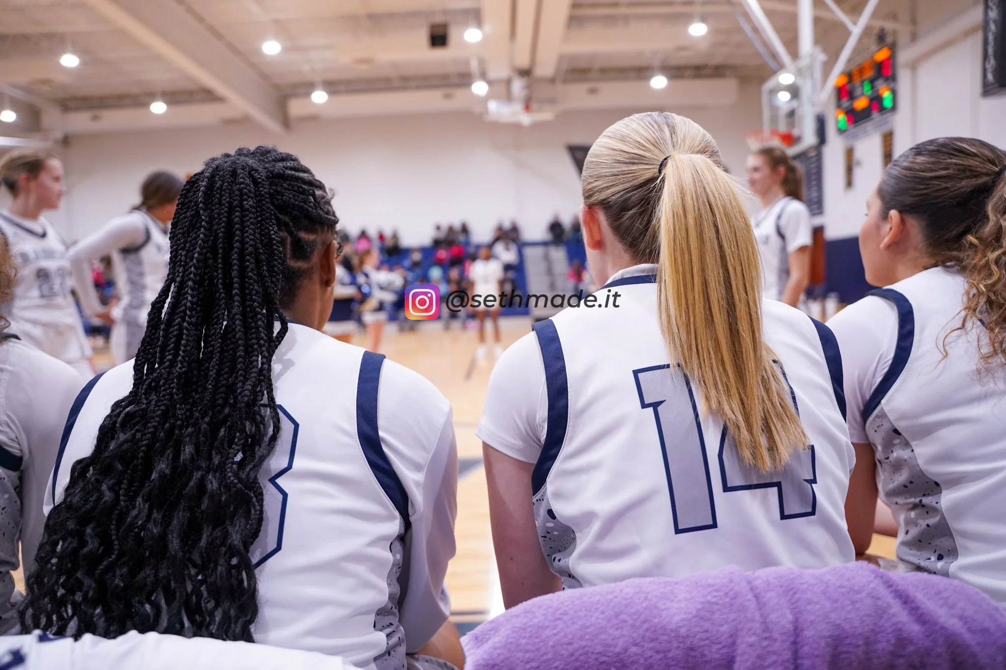 Women basketball players sitting on the bench during a game, wearing white jerseys with blue numbers, inside a gymnasium.