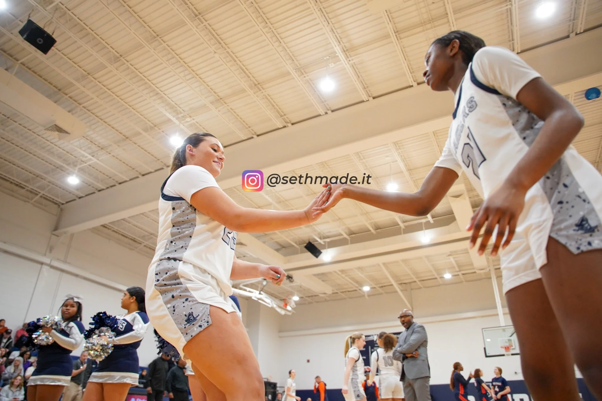 Two female basketball players are exchanging high fives during a game, with cheerleaders and spectators in the background.