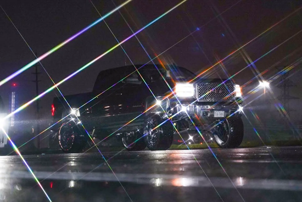 A black truck reflecting bright lights at night on a wet road, with starburst effects and lens flare creating colorful streaks across the image.