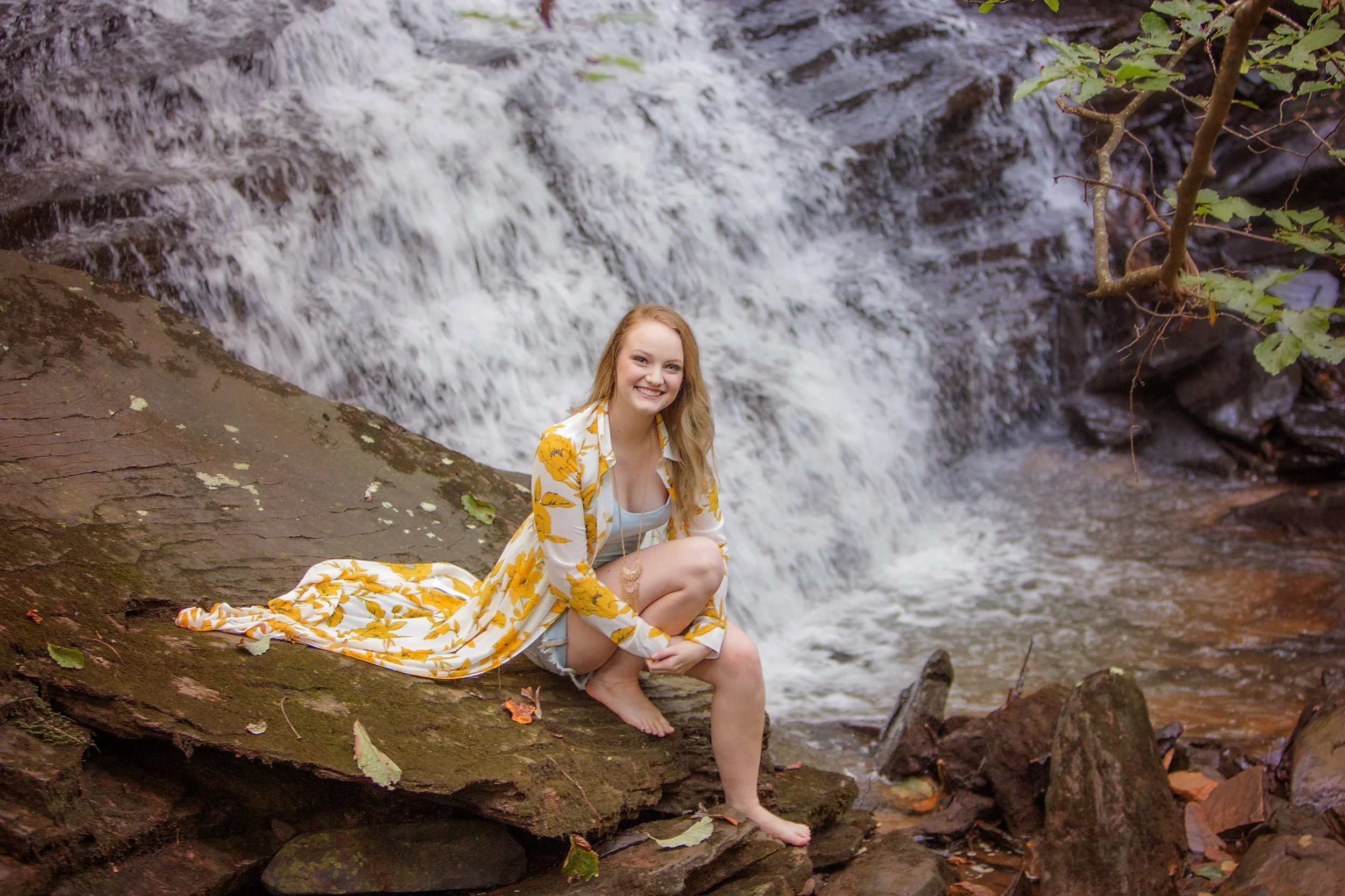 high school senior sitting on rock by waterfall smiling