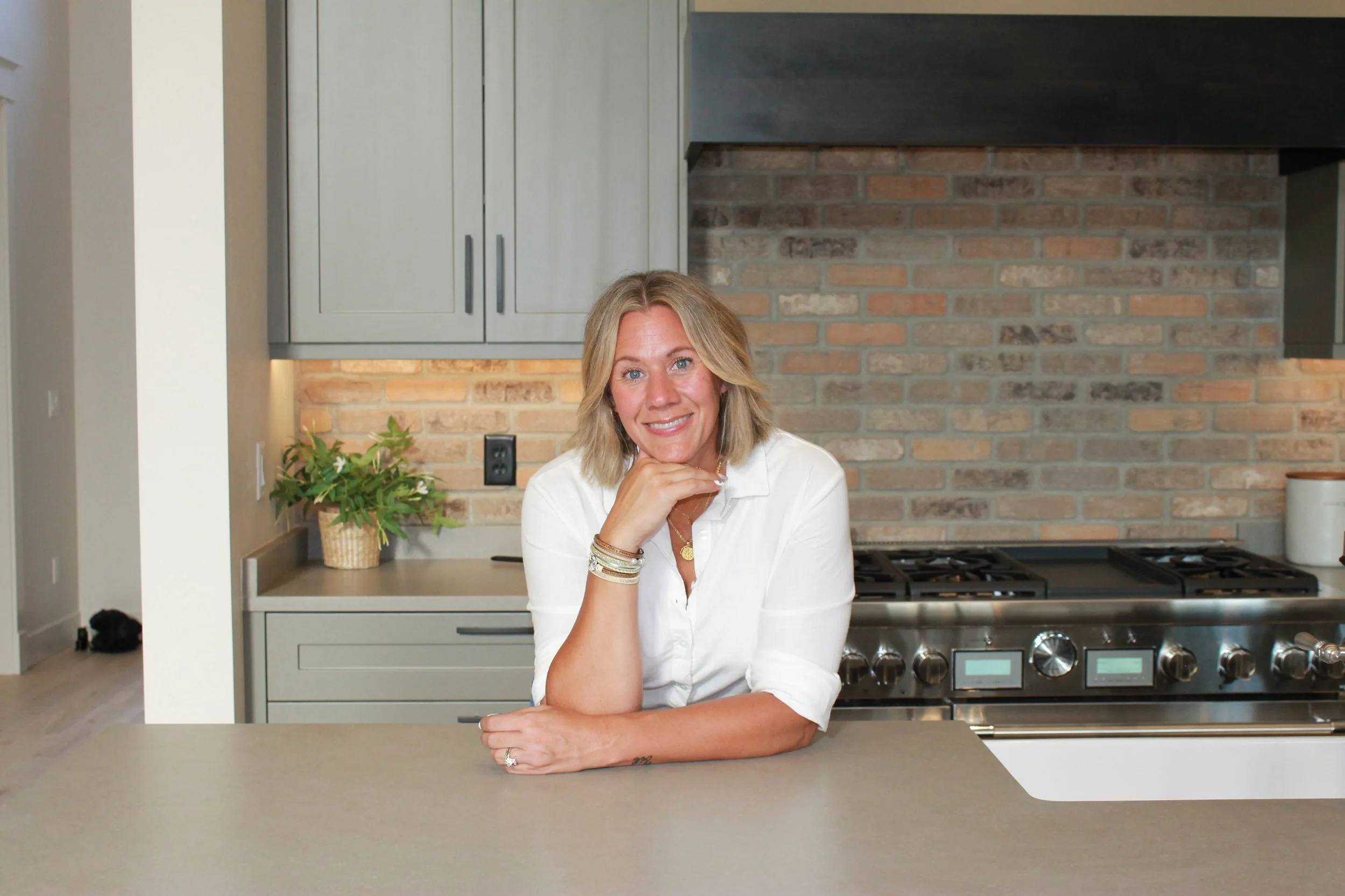A woman with blonde hair, wearing a white shirt, sitting at a kitchen counter with her right elbow resting on the surface. She is smiling and looking directly at the camera. The background features a brick wall, gray cabinetry, a plant in a basket, and a stove.