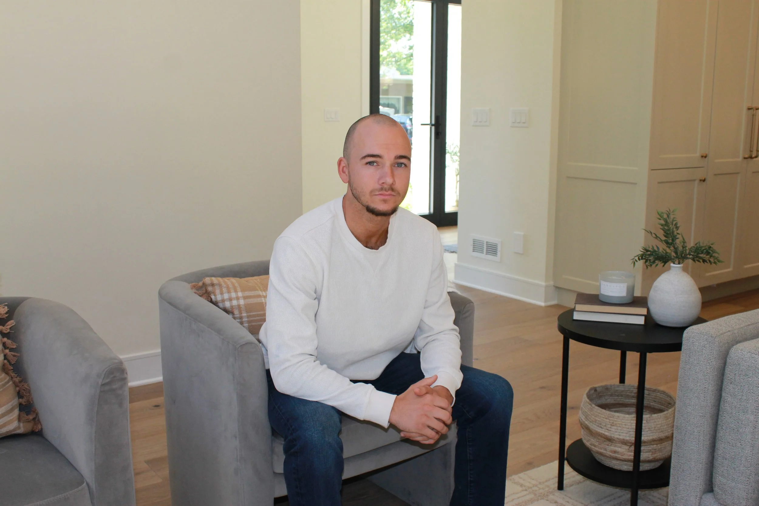 A young man with a shaved head and facial hair sitting on a gray armchair in a bright living room with hardwood floors, a door with glass panels in the background, and a side table with books, a plant, and a candle.