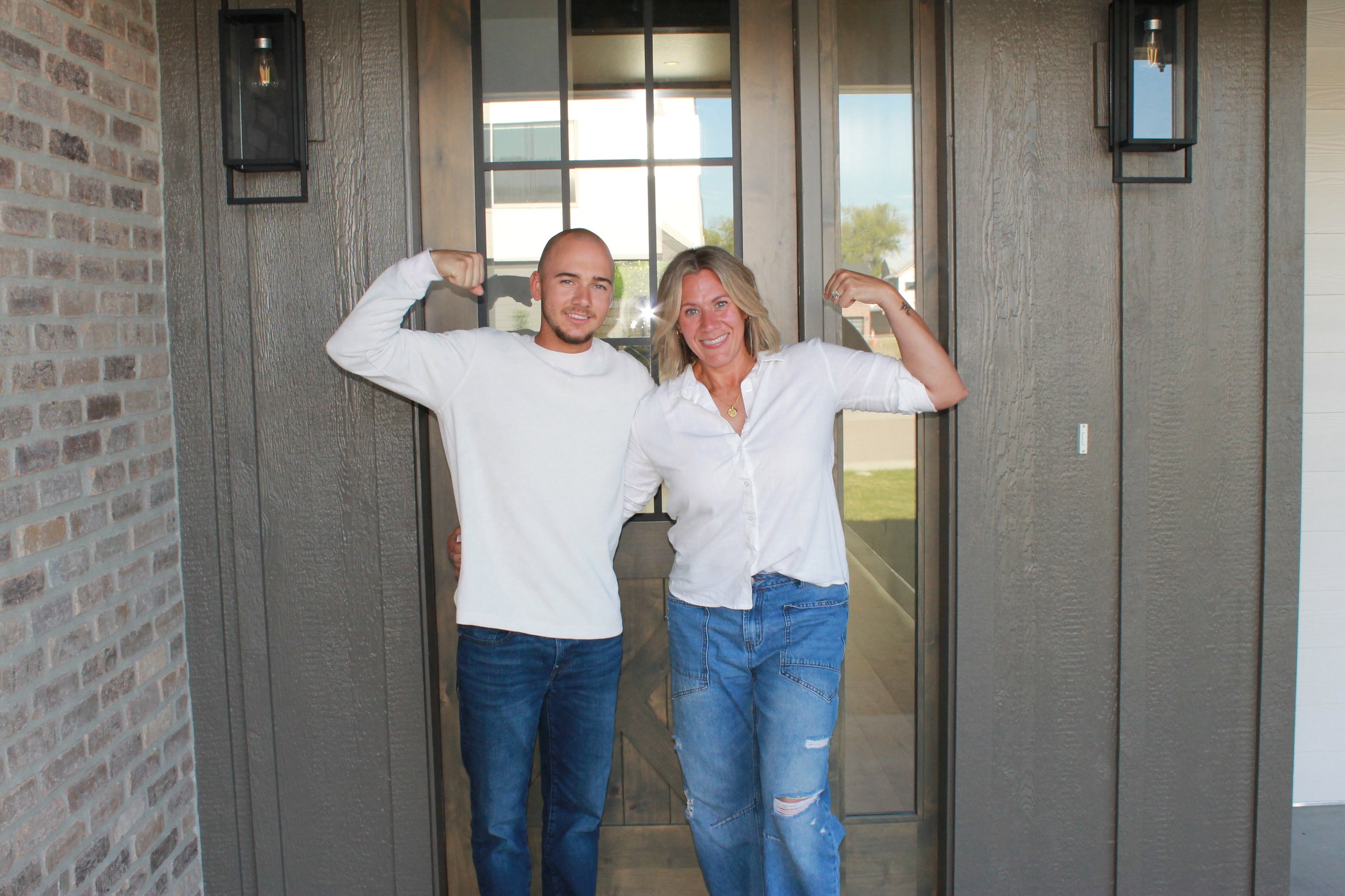 A young man and a woman stand side by side at the front door of a house, flexing their biceps and smiling. The woman has blonde hair and is wearing a white shirt and ripped jeans, while the man has a shaved head and is wearing a white long-sleeve shirt and jeans.