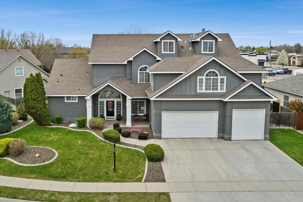 A gray, multi-story suburban house with attached two-car garage, landscaped front yard with lawn, bushes, and trees, and a sidewalk in front.
